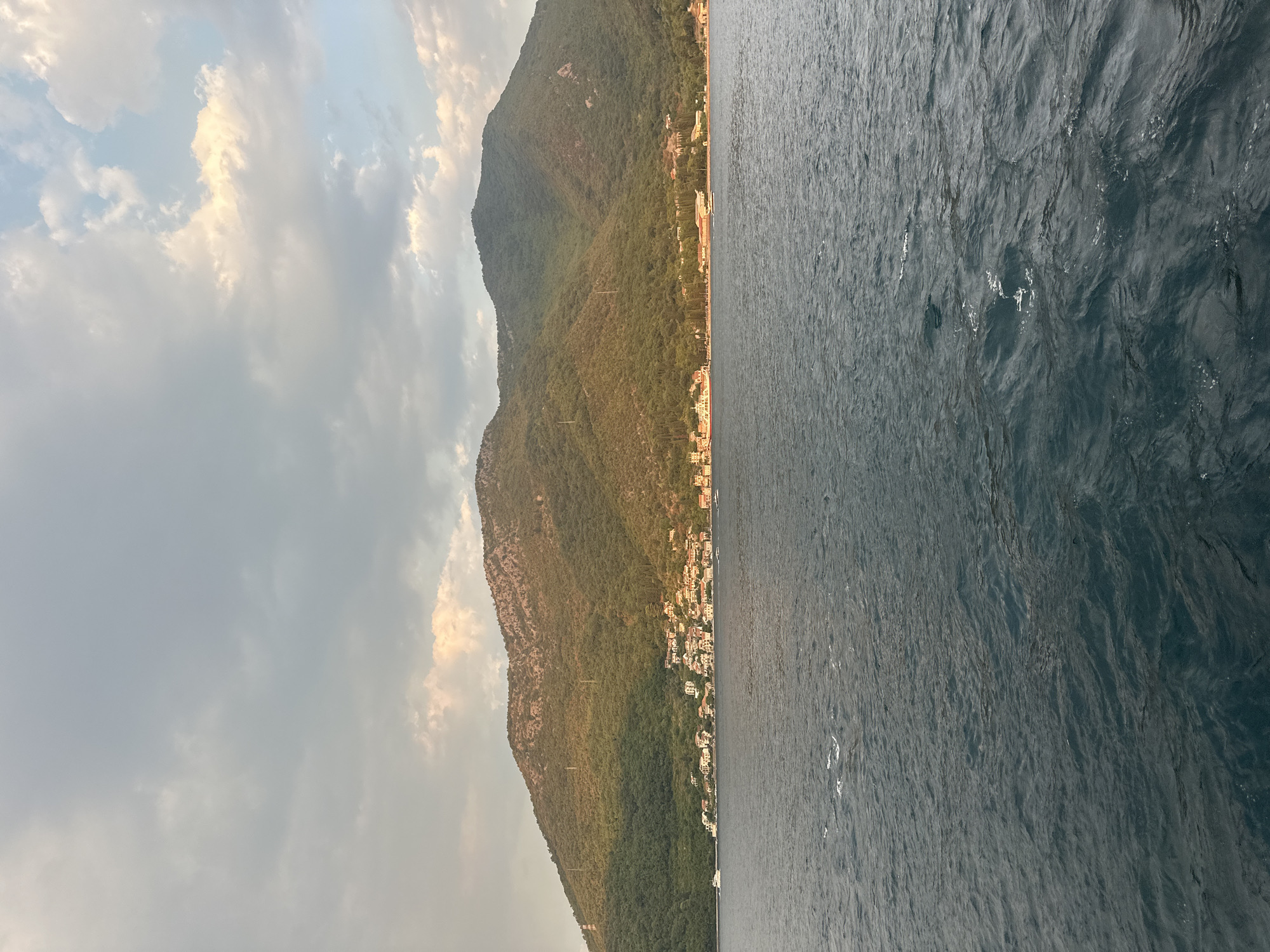 The image depicts a scenic view of a coastal town nestled at the base of a lush, green mountain range. The water in the foreground is a deep blue, with gentle ripples indicating calm conditions. A small white boat can be seen on the left side of the frame, moving towards the shore. The town is characterized by its white-washed buildings with red-tiled roofs, clustered along the coastline. Trees and vegetation cover the mountain slopes, with some areas appearing more rugged and rocky. The sky above is a mix of gray clouds and patches of blue, with sunlight filtering through, casting a warm glow on the mountainside. The overall impression is one of tranquility and natural beauty. Based on the architectural style and the mountainous coastal landscape, the location is likely a town in the Bay of Kotor, Montenegro, possibly resembling areas around Đurići.