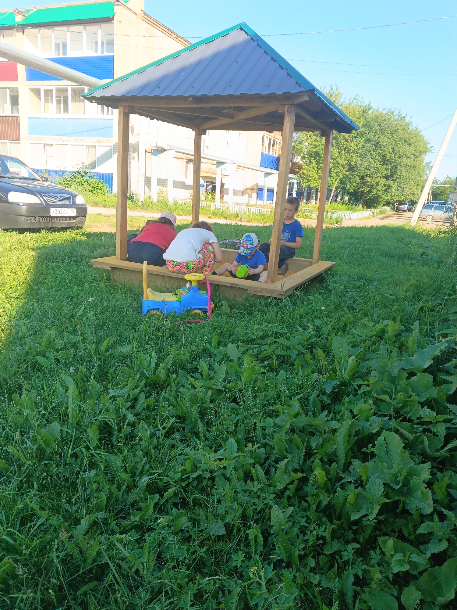 In this outdoor daytime scene, likely in Tolbazy, Russia, a wooden gazebo with a dark blue metal roof stands over a sandbox. Three children are present: two girls and one boy. The two girls are seated in the sandbox, one wearing a red shirt and the other a white shirt with brightly patterned leggings. The boy, wearing a blue t-shirt and a baseball cap, is also in the sandbox, holding a green watering can. A blue toy truck with a yellow handle is situated near the children in the sandbox.
In the background, a multi-story building with blue and beige siding is visible, along with a black car parked on the left side of the frame, displaying the license plate "x302cx 102". The area surrounding the sandbox is covered in lush green grass and various plants. Trees line the background, and the sky above is clear blue, suggesting fair weather. The overall atmosphere appears relaxed and leisurely, indicative of children playing outdoors on a pleasant day.