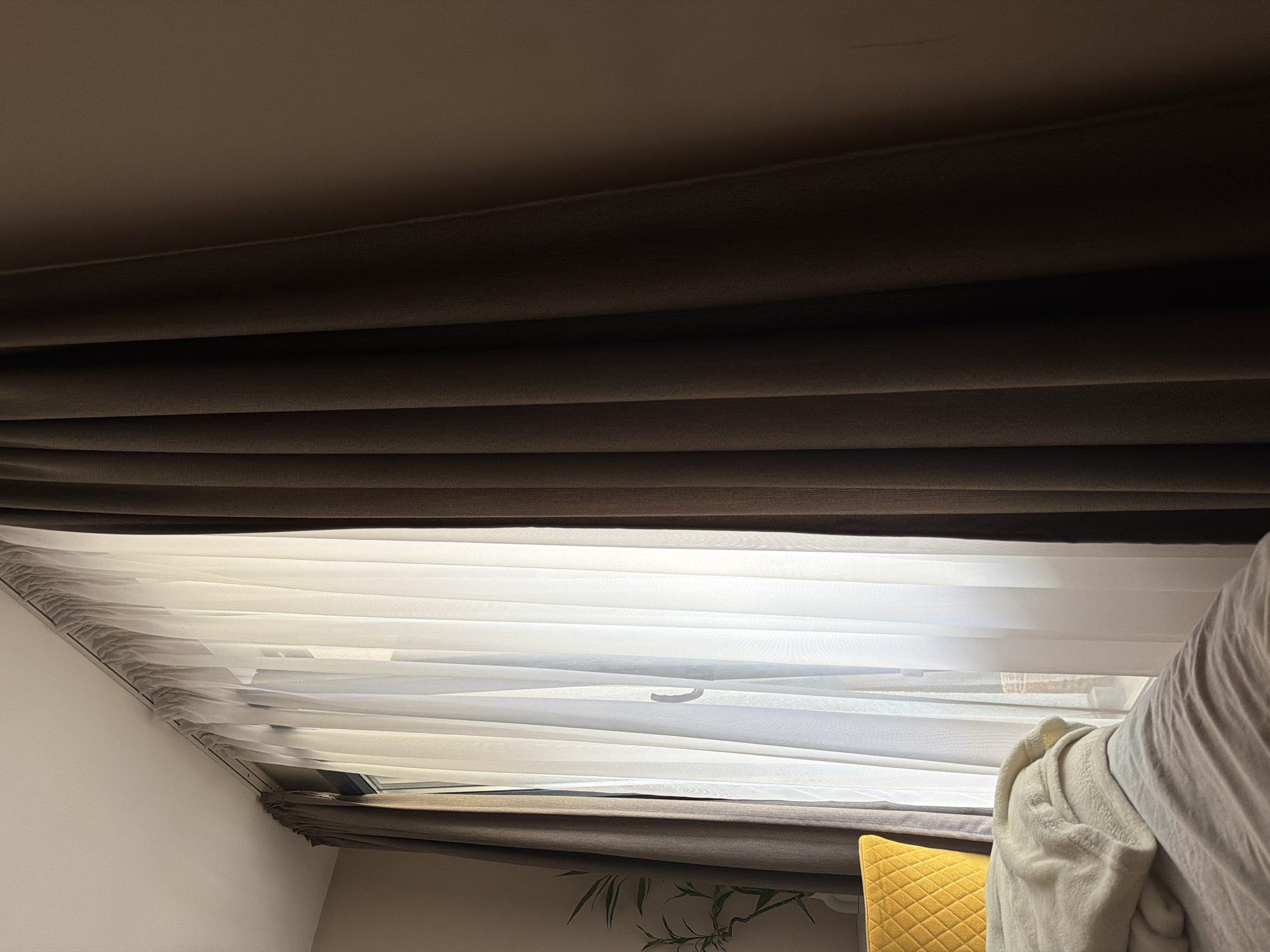 The image depicts an indoor scene, likely a bedroom or hotel room, illuminated by natural light entering through a large window.
The central and upper portions of the frame are dominated by a window covered with a double layer of curtains. The inner layer consists of sheer, white, vertically pleated fabric, partially drawn to reveal a portion of the window with a visible white handle. The outer layer comprises heavy, dark brown, vertically pleated fabric. A section of the dark brown curtain is visible on the far left, partially drawn. A significant portion of the same dark brown curtain occupies the entire right side of the image, running from the ceiling to the bottom edge, obscuring a light brown wall. All curtains are suspended from a white, ceiling-mounted track system.
In the lower foreground, a rumpled bed is visible, covered with light-colored bedding. A folded, light-colored, textured blanket rests on the bed. To the left of the bed, a segment of a yellow, quilted furniture piece, possibly a chair or headboard, is partially visible. Further left, behind the yellow furniture, a green plant with multiple stems and leaves stands in a white vase, positioned against a light brown wall. The ceiling is white.
No people or active interactions are depicted. The environment suggests a calm, private space. The image provides no visual clues to specify a location within the city of Sofia, Bulgaria.