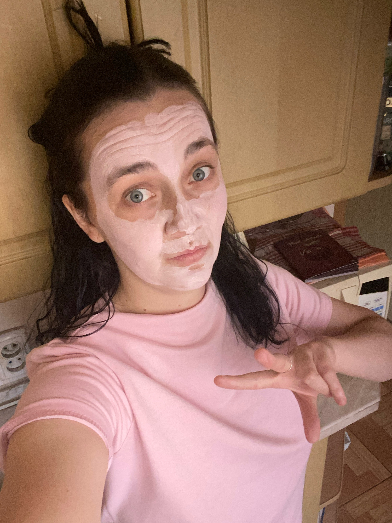 An overhead, medium shot shows a young woman with dark hair pulled up in a messy bun, wearing a pink t-shirt. She is applying a pink face mask to her face. Her eyes are wide and blue, and she is looking directly at the camera. Her right hand is raised and gesturing towards the camera. The background appears to be a kitchen with wooden cabinets and countertops.