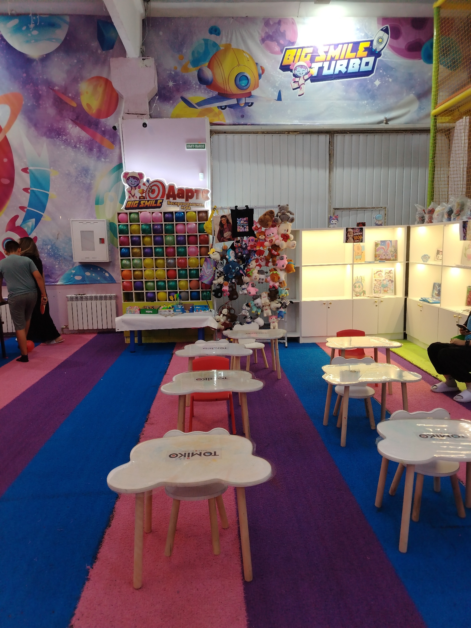 The image depicts the interior of an indoor amusement park or entertainment center. The scene is colorful, with a brightly painted wall featuring a space-themed mural and a large banner displaying "BIG SMILE TURBO." In the foreground, there are several small, white, cloud-shaped tables with wooden legs, set up on a carpeted floor divided into sections of pink, blue, and purple.
In the middle ground, there is a game stall with a grid of colorful balloons, presumably for a dart-throwing game. Behind this, a white shelving unit is stocked with various stuffed animals and toys. A few people are visible; a young boy in a grey t-shirt and shorts is standing near the game stall, facing away from the camera. A person in a black outfit is partially visible behind him. Further back, near the shelves, a person is seated on the floor, wearing black pants and white sandals.
The environment suggests a children's entertainment venue, likely a place for parties or casual play. The overall impression is one of a fun and stimulating atmosphere designed for children. The location of this amusement park is identified as being in the city of Taldykorgan, Kazakhstan.