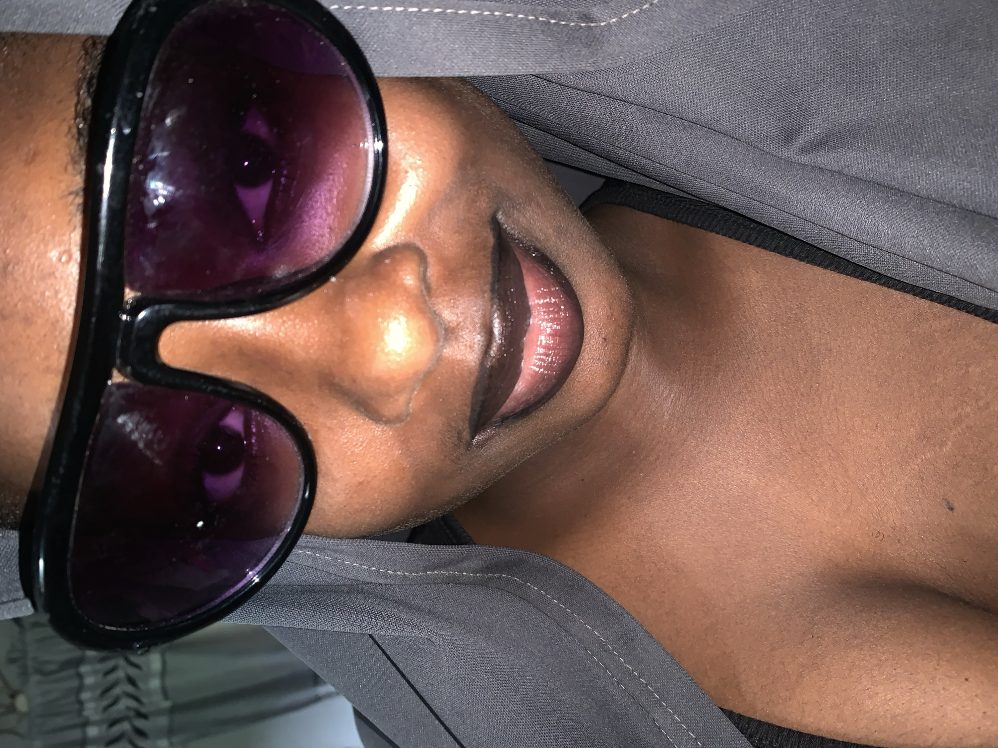 A close-up, head-and-shoulders shot captures a person with dark skin wearing large, black-rimmed sunglasses with purple lenses. The individual's lips are painted with dark, glossy lipstick. They are wearing a dark grey hooded garment that partially obscures their face and shoulders. The background is out of focus, showing what appears to be a patterned curtain on the left side. The lighting is dim, creating shadows and highlighting the sheen on the person's skin and lips. This image was taken in Rafin Par, Nigeria.