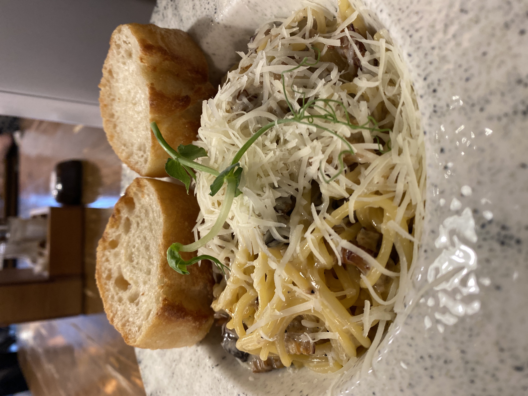 The image shows a plate of pasta next to two slices of bread. The pasta is covered in grated cheese and garnished with a small sprig of greens. The plate has a speckled pattern. In the background, there are blurry glimpses of a table and other objects. The location is Yekaterinburg, Russia.