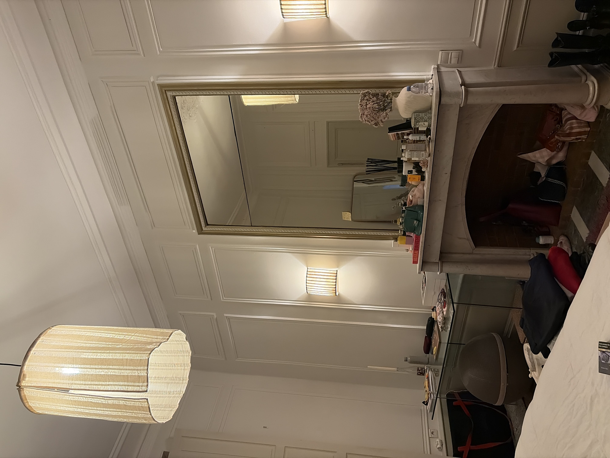A residential interior in Paris, France, is depicted, characterized by classical Haussmannian architectural elements. The room features high white ceilings with crown molding and white walls with extensive paneling (boiseries). Artificial illumination is provided by a central ceiling pendant lamp with a light-colored, vertically striped fabric shade, and two matching fabric-shaded wall sconces.
A large, ornate gold-framed mirror dominates the right side of the frame, positioned above a white marble fireplace mantelpiece. The fireplace opening, or hearth, is utilized for storage, containing multiple bags, including a red tote, a black quilted chain-strap bag, a striped fabric bag, and a pale pink bag. The mantelpiece surface is densely arranged with personal effects: a white ceramic vase holding dried light-colored flowers, a clear water bottle, various beauty products, perfumes, a small teal handbag, a smaller gold-framed rectangular mirror, a reed diffuser, and several books or magazines.
To the left, a transparent glass-top console table with thin legs is positioned against the wall, holding books, papers, and a white water bottle. Beneath this table rests a dark grey, egg-shaped pet bed or furniture piece, alongside a large black tote bag with red handles. In the immediate foreground, the edge of a bed with white linens is visible. A stack of folded clothing, including dark blue and red garments, is located on or near the bed. On the floor to the right, adjacent to the fireplace, several pairs of dark shoes and tall black boots are arranged. No individuals are present in the scene.