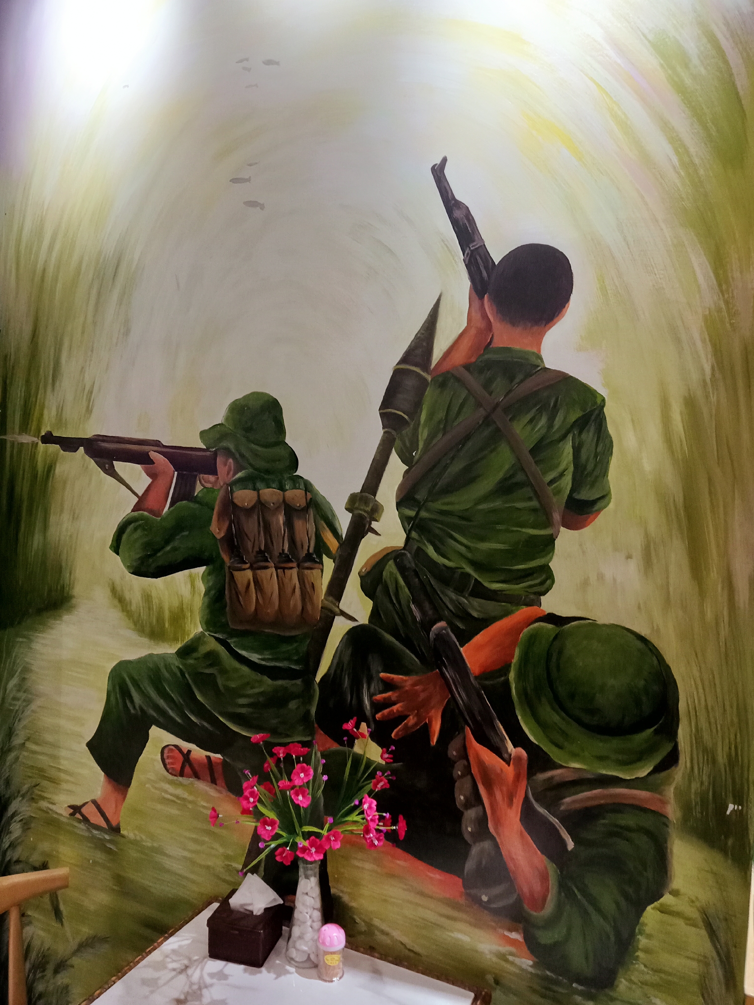 This image presents a vibrant painting, likely a mural, depicting three soldiers in a dense, green, jungle-like environment. The perspective places the viewer behind and slightly above the figures, who are depicted in various states of readiness.
The central figure, viewed from the back, wears a short-sleeved green uniform and carries a rocket-propelled grenade (RPG) launcher on his left shoulder, pointing upwards. His right hand appears to be holding a rifle, also pointed towards the upper right. To his left, another soldier, seen from the back in a crouched position, wears a long-sleeved green uniform, a wide-brimmed hat, and a brown canvas backpack. He is actively aiming a rifle forward, with a small muzzle flash indicating a shot has just been fired or is about to be. The third figure is partially visible in the bottom right foreground, also in a green uniform and hat, holding a weapon. The soldiers are clad in simple green fatigues; the soldier on the left wears sandals.
The background features sweeping brushstrokes of green and yellow, suggestive of tall grass or dense foliage forming a natural pathway or tunnel, leading towards a bright, luminous area at the top of the painting, implying daytime. Faint, dark, elongated shapes, possibly birds or distant aircraft, are discernible in the bright upper-left background.
In the immediate foreground, real-world objects are visible on a white table: a bouquet of bright pink flowers in a clear vase, a dark brown tissue box, and a small pink-capped container (possibly a condiment shaker). These items indicate the painting is a wall mural within an interior setting, likely a restaurant or home in Nha Trang Ward, Vietnam, given the provided context. The painting evokes a sense of military readiness or engagement in a tropical combat zone. There is no visible text in the painting or on the foreground objects.