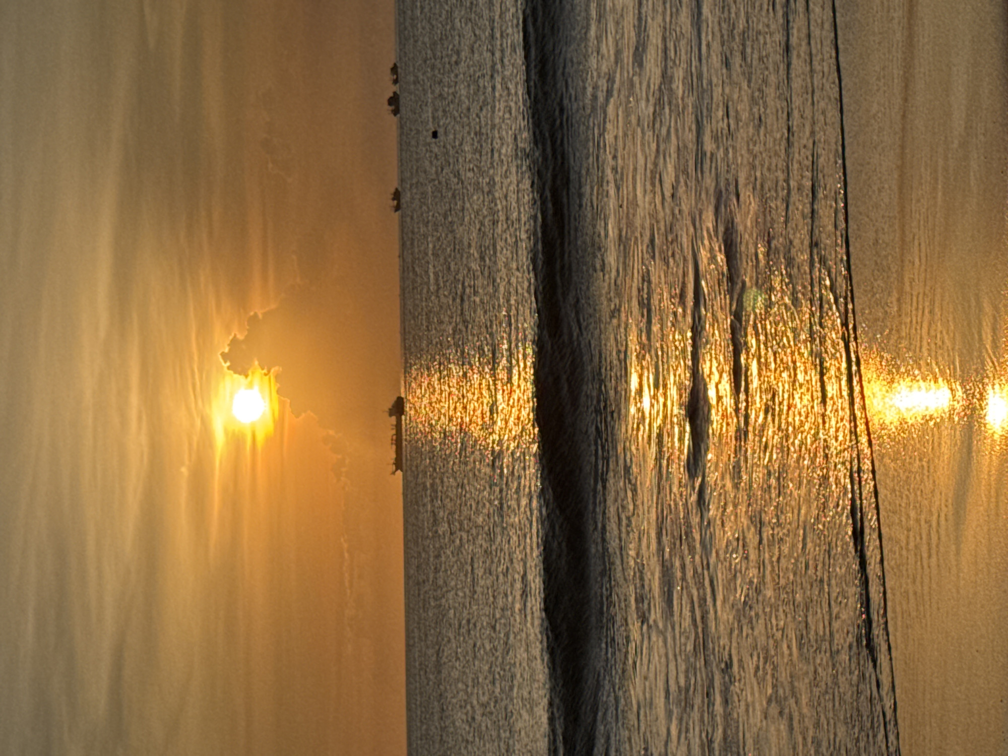 The image shows a sunset over the ocean, likely in Kuta, Indonesia. A bright sun is partially obscured by clouds, casting a golden glow across the sky and water. The ocean surface reflects the sunlight, creating shimmering patterns. A pier or jetty is visible in the water. Birds are perched on the wooden post.