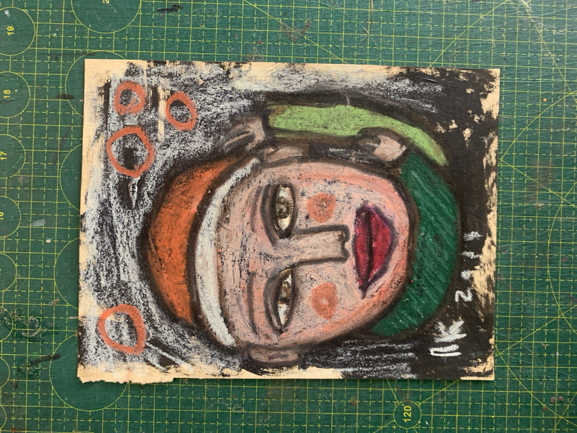 The image displays a colorful, abstract portrait of a person's face, rendered in a textured style with oil pastels on a rough, beige paper. The face is predominantly pink and peachy, with large, expressive eyes and red lips. The person is wearing an orange hat and a green scarf. The background is a mix of black, grey, and white scribbles. In the upper portion of the image, there are several incomplete orange circles. The lower section of the paper contains a signature or inscription in white, appearing to be "RK 2014". The artwork is placed on a green cutting mat with a grid pattern, which is common in art studios or craft areas. There is no indication of Sergeevo, Russia in the image.
