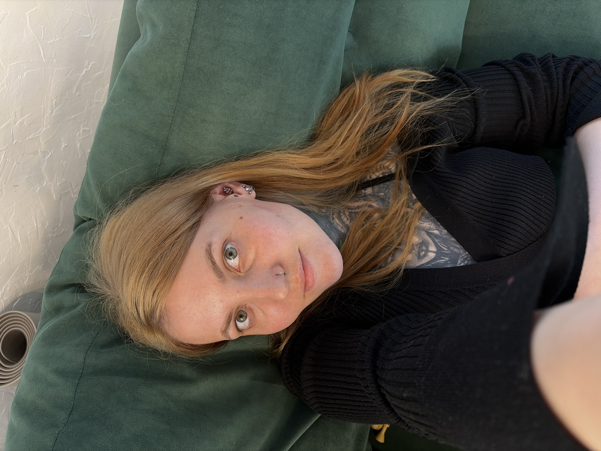 A woman with fair skin and light brown hair is lying on a green, cushioned surface, likely a sofa or a large cushion, indoors. She is wearing a black ribbed top and has a tattoo visible on her chest. She has multiple piercings on her left ear and a nose ring on her left nostril. A rolled-up yoga mat is visible in the background to her left. The setting appears to be a residential interior. There is no indication of the location being in Malyi Bukor, Russia.