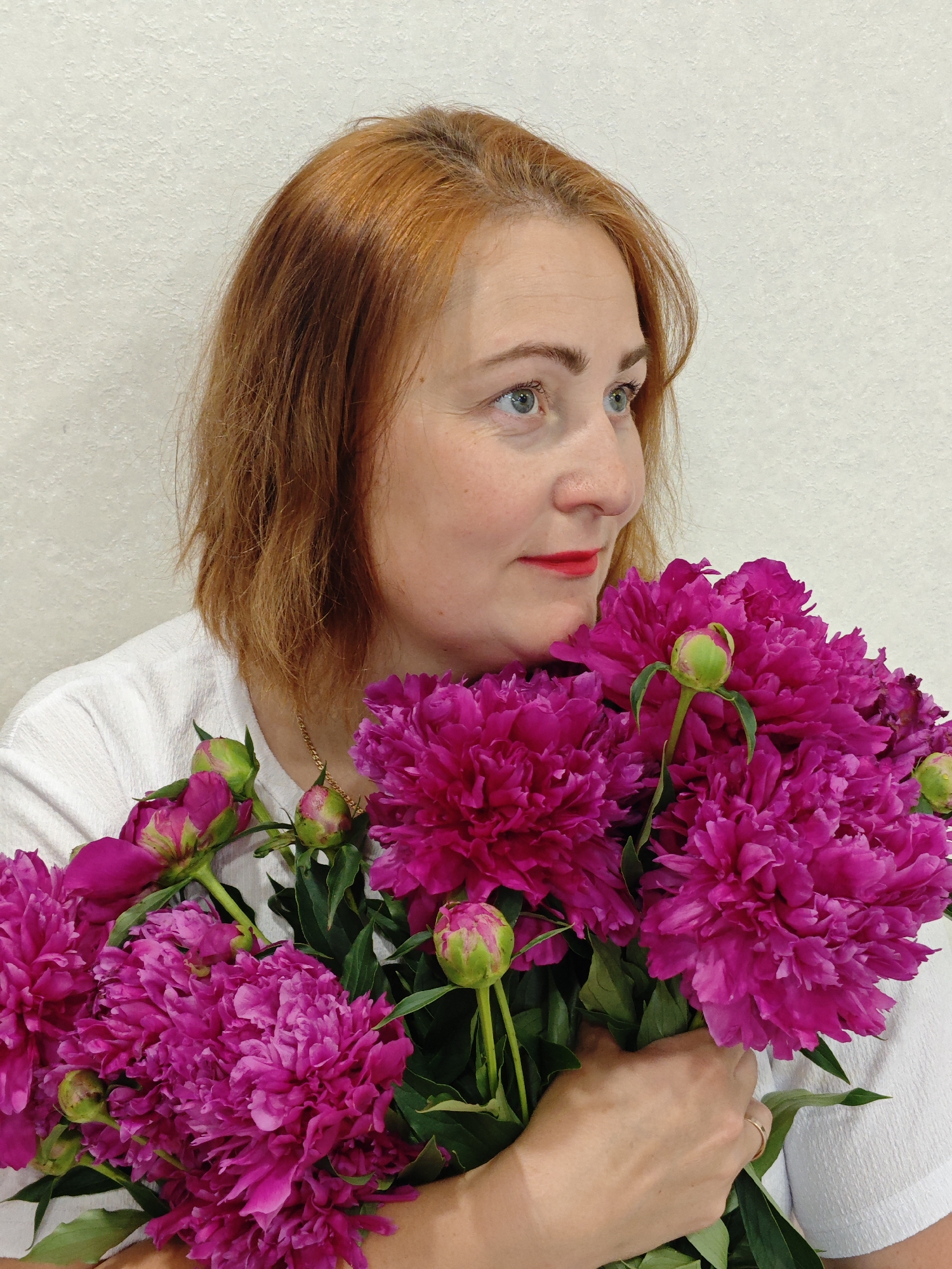 A woman with auburn hair and fair skin, wearing a white shirt, holds a bouquet of bright pink peonies. She has blue eyes and red lipstick. The background is a plain, textured off-white wall. The scene is indoors, likely a home, and the lighting is bright. There is no indication of the city of Markovskii, Russia, in the image itself.