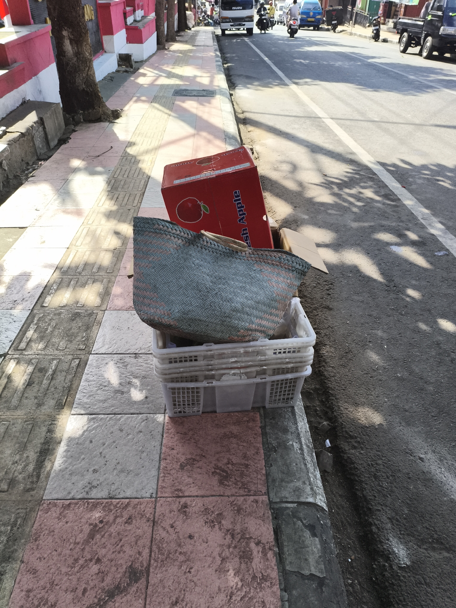 On a sunny day in Magelang, Indonesia, a roadside scene unfolds. A red cardboard box, labeled "Fresh Apple" and featuring a graphic of an apple, sits beside a woven basket filled with what appears to be plastic bags. These items are placed on top of two stacked white plastic crates on a patterned sidewalk made of pink, gray, and white tiles. A tree with a rough bark trunk grows from the sidewalk, its base partially obscured by a concrete structure. The sidewalk is adjacent to a paved road where various vehicles are visible, including a white truck with "ISUZU" written on it, a blue car, and several motorcycles. Shadows from overhead foliage are cast across the sidewalk and the road, indicating a tree-lined street. The overall environment suggests a commercial or residential area in the city.