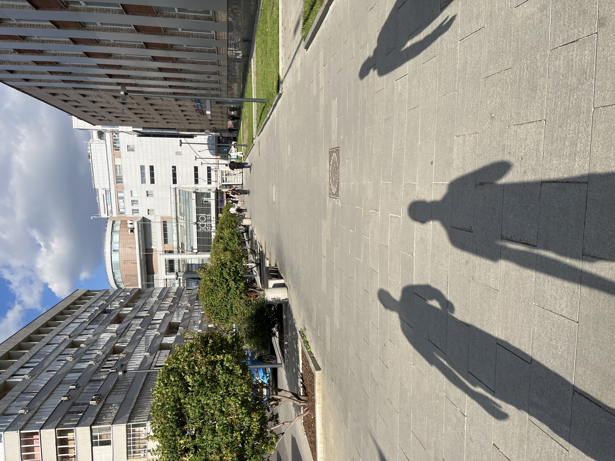 This media file depicts a sunlit pedestrian walkway bordered by modern buildings and trees. The scene is captured from a slightly elevated, downward-angled perspective, and the image is rotated 90 degrees clockwise. Shadows of people are cast on the stone-tiled pavement, indicating a sunny day.
In the foreground, several long, distinct shadows of people are visible on the ground, stretching towards the viewer. These shadows suggest people are walking away from the camera or standing in the direction of the sun. Further down the walkway, a few small groups of people are walking.
On the left side of the walkway, there are trees planted in planters. Beyond the trees, a series of modern buildings are visible. The building closest to the foreground on the left is a multi-story apartment building with a repetitive facade of windows. Further back, a white building with many windows and a curved section is visible, and behind that, another building with a brick and metal facade. On the right side of the walkway, beyond the shadows, more of the paved area is seen, leading towards the buildings on that side. A circular metal grate is embedded in the pavement.
The time of day appears to be late morning or early afternoon, judging by the length and direction of the shadows. The weather is clear and sunny, with some clouds visible in the sky above the buildings on the left.
There is some visible text on signs attached to the buildings. On the white building in the middle, "lido dress$" is visible. On a blue car parked in the distance, part of "Яндекс" (Yandex) can be seen.
The overall impression is an urban scene on a pleasant day, with people enjoying a walk on a paved path in a city environment. The location cues, such as the architecture and the visible text, suggest Moscow, Russia.