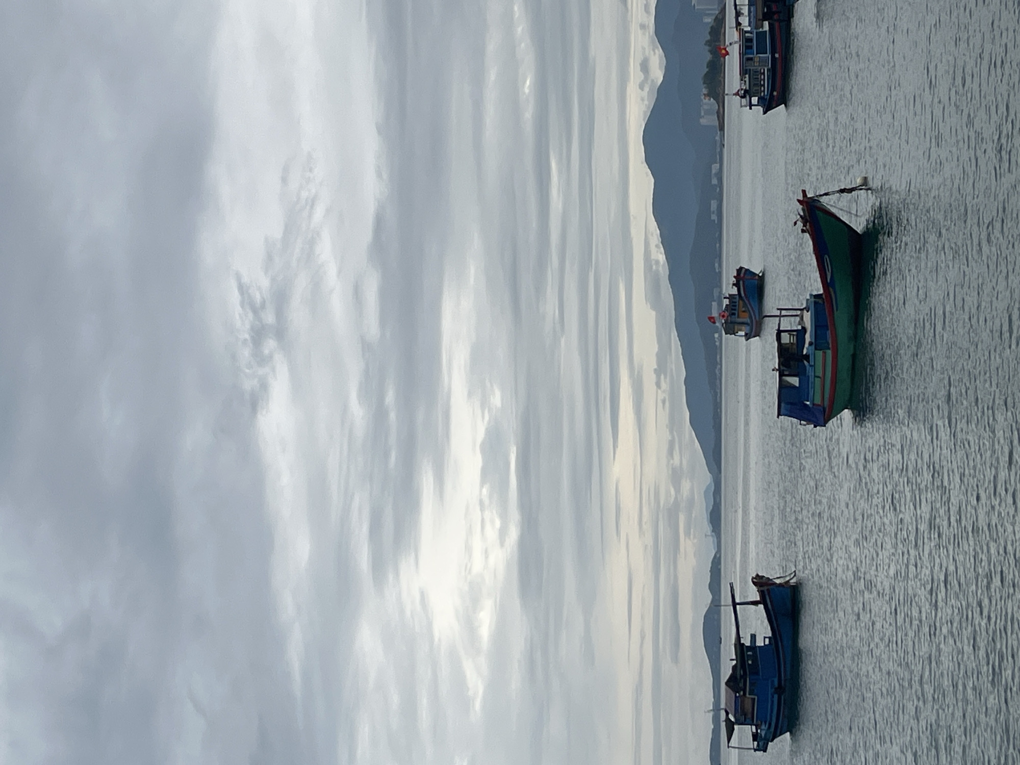 The image captures a scene on the water in Bac Nha Trang Ward, Vietnam, under a cloudy sky. Several fishing boats are visible on the water, with mountains in the background. The boats are colorful and varied in size. The water appears calm with visible ripples. The overall atmosphere is subdued due to the overcast sky.