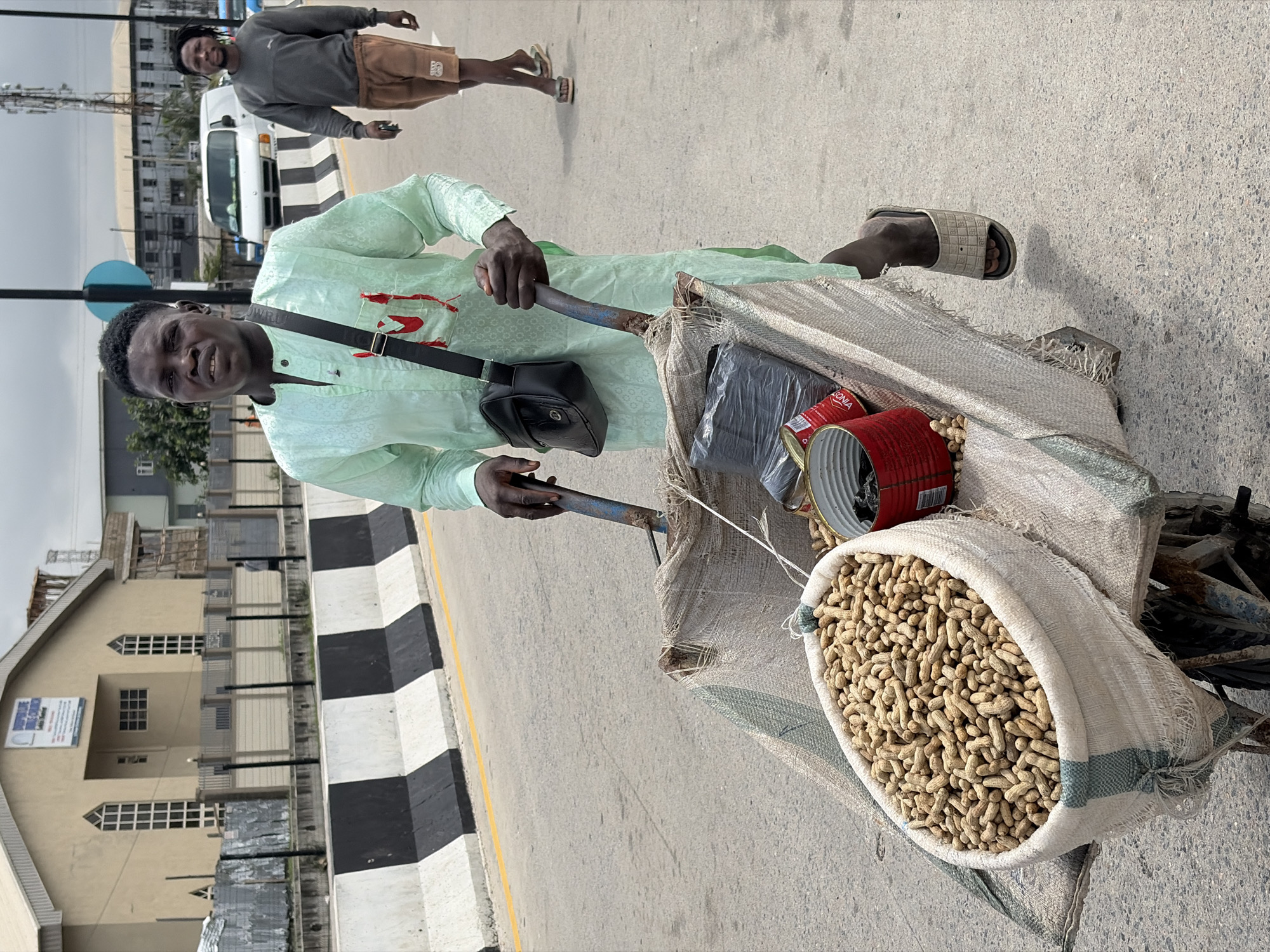 In Igboefon, Nigeria, a man wearing a light green traditional outfit and a black crossbody bag pushes a two-wheeled cart filled with goods. The cart's large front container is packed with raw peanuts in their shells. Behind the peanuts, several red and silver cans are visible, along with a black plastic-wrapped item. The man's hands grip the cart's handles, and he wears a light-colored sandal on his right foot. Behind him, another man in a gray long-sleeved shirt and brown shorts walks away from the camera, also wearing sandals. They are on a paved road bordered by a black and white striped barrier. In the background, buildings, a white van, and a cell tower are visible under an overcast sky.