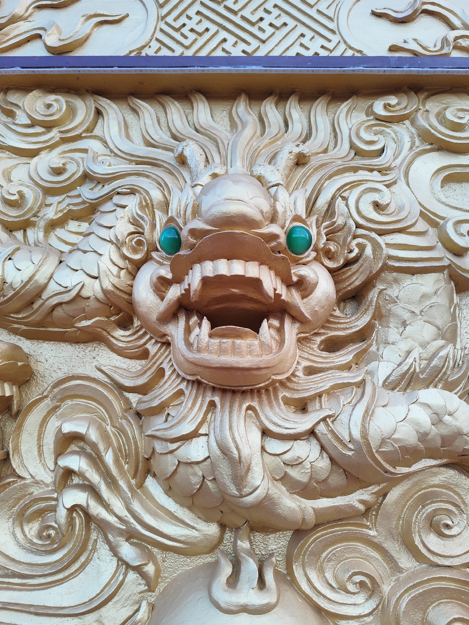 This close-up image showcases a vibrant golden relief sculpture of a dragon's head and upper body, rendered in a traditional East Asian style. The primary subject is the dragon, depicted with intricate details, an open mouth revealing sharp teeth, and striking emerald-green, spherical eyes. Its head is surrounded by flowing, stylized mane-like elements, while scaled portions of its body are visible on either side.
The dragon is set against a textured background of swirling, cloud-like patterns, also rendered in gold, which create a sense of movement and depth. Above the dragon's head, at the very top of the image, is a horizontal strip of wood or stone with a repeating geometric, labyrinthine pattern. Below this patterned strip and above the main dragon relief, there is a narrow, darker horizontal band, possibly an architectural element, which appears to have a series of small, embedded lights, creating subtle reflections.
The sculpture's golden color is prominent, with varying shades and reflections indicating bright, likely daytime lighting conditions. The overall impression is one of power and grandeur, characteristic of decorative elements found in temples or significant buildings in cultures like Vietnam, aligning with the given location context of Lạc Dương. No visible text is present. The detailed carving and metallic sheen highlight the craftsmanship of the piece.