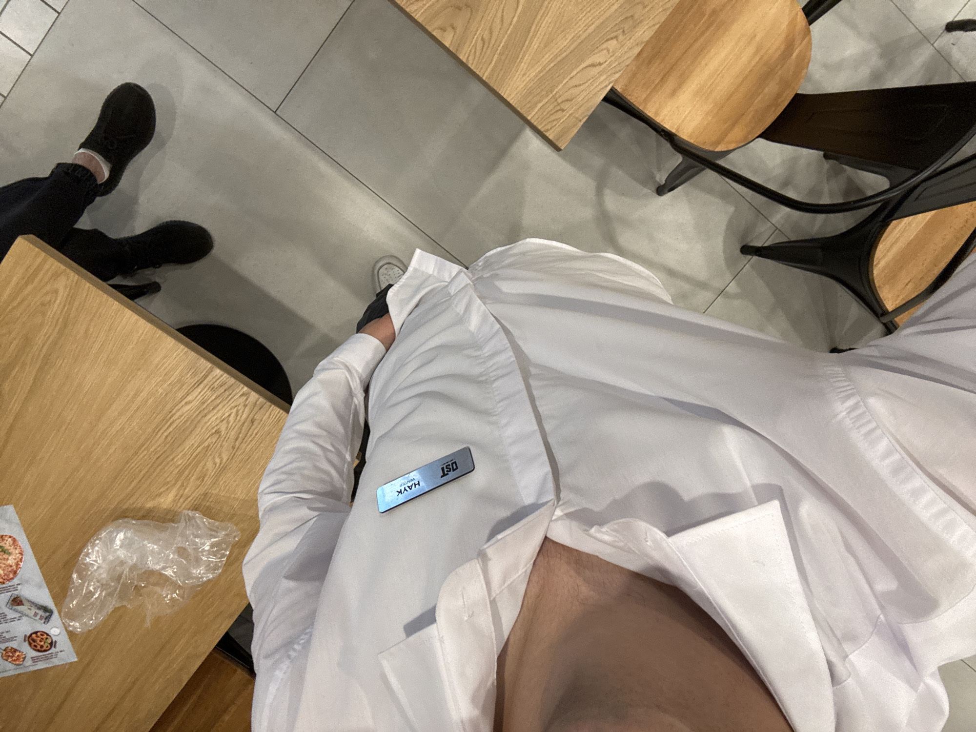 The image presents a first-person, overhead perspective. A person wearing a white collared shirt with a name tag that reads "HAYK WAITER ISD" is positioned near wooden tables. On one table, there is a pizza menu and a clear plastic bag. The person is seated at a table with wooden chairs. The setting suggests an indoor dining area, possibly in Yerevan, Armenia, given the name "HAYK," which is a common Armenian name. Another person is visible in the corner of the frame, wearing black shoes. The environment appears to be a restaurant or cafe, with a focus on the individual wearing the white shirt.