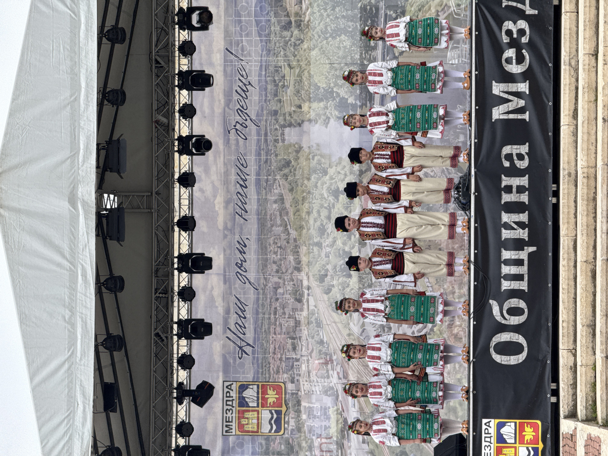The image shows a scene in Mezdra, Bulgaria, likely a cultural event or performance. A group of children dressed in traditional Bulgarian folk costumes stand in a line, possibly preparing to dance or perform. Behind them is a backdrop with the words "Наши дом, наше сърце!" (Our home, our heart!) in Bulgarian, as well as the city's coat of arms and the word "МЕЗДРА" (Mezdra). To the left of the backdrop, there's a stage structure with lighting equipment. To the right, a black banner reads "Община Мездра" (Municipality of Mezdra), along with the city's coat of arms again. The scene is set in an outdoor location with stone steps visible on the right side.