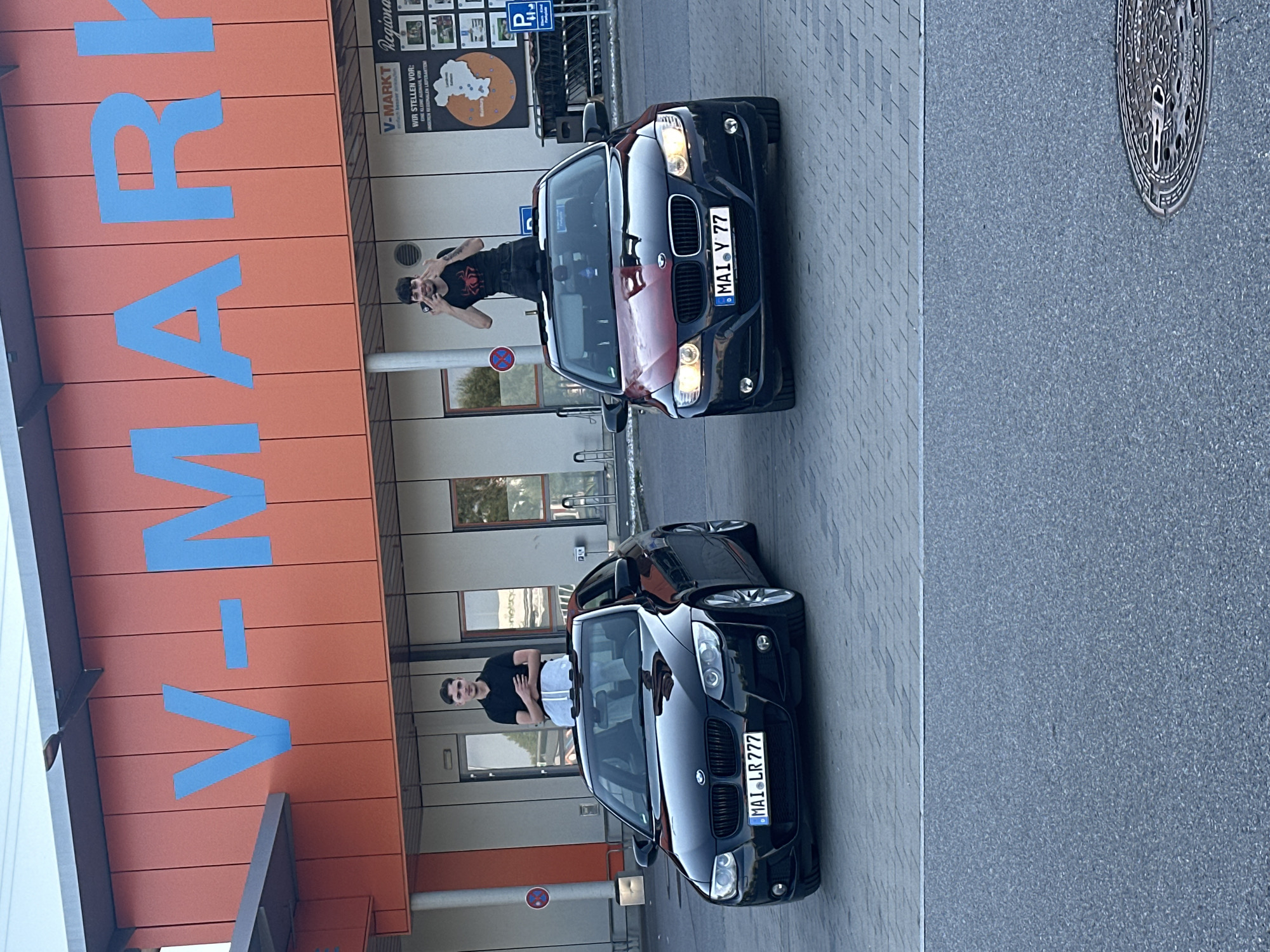 The image displays two young men posing through the open sunroofs of two black BMW cars in a parking lot in Mainburg, Germany. In the background, a V-MARKT supermarket building with a large orange facade featuring prominent blue "V-MARKT" lettering is visible.
The man on the left, standing through the sunroof of a black BMW coupe (license plate: MAI LR 777), is wearing a black polo shirt and light-colored shorts, with his arms crossed over his chest. He has short dark hair and is looking towards the camera.
The man on the right, standing through the sunroof of a black BMW sedan (license plate: MAI Y 77), is wearing a black t-shirt with a red spider graphic and is making a peace sign with both hands. This car's hood appears to be split, with the right half being a contrasting reddish-brown or burgundy color against the black body. He has short dark hair and some facial hair.
The parking lot surface directly beneath the cars consists of light grey interlocking pavers, transitioning to a darker grey asphalt surface in the foreground, where a circular metal manhole cover is visible in the lower right corner. The supermarket building's lower section features lighter panels, windows, and doors, with a pillar displaying a red "no stopping" sign. Another blue sign for "P Kundenparkplatz" (Customer Parking) is visible on the far right. The scene is illuminated by bright, natural daylight.