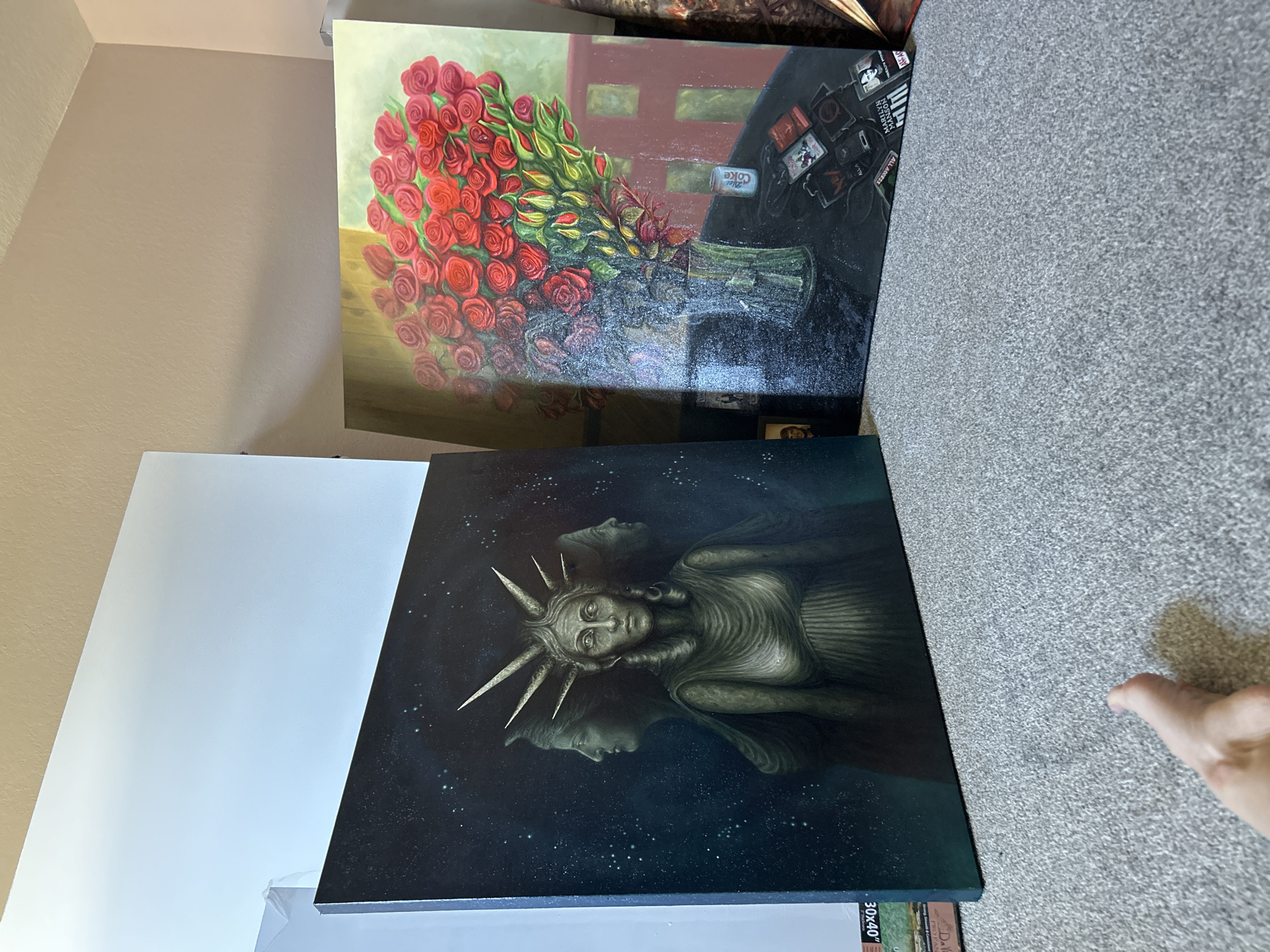 Two large artworks are positioned indoors, leaning against a light-colored wall and resting on a grey carpet. The image appears to be rotated 90 degrees clockwise from its natural orientation.
The top painting features a vibrant still life with a large, lush bouquet of red roses, complete with green stems and leaves, placed in a green glass vase. This arrangement sits on a dark surface, likely a table, with a reddish-brown background resembling an interior wall or window frame. Next to the vase on the table are various small objects: a can of Diet Coke, and several rectangular items resembling album covers or concert memorabilia. Visible text on these items includes "DIET COKE", "MARILYN MANSON", and "ALL AGES".
The bottom painting depicts a surreal, dark-toned scene. The central figure is a stylized, somewhat emaciated representation of the Statue of Liberty, identifiable by her spiked crown and draped form. Her face is pale and her eyes are light, creating a stark contrast against the dark, starry blue and black background. Below and behind her, faint reflections or ghostly secondary faces of the figure are visible. Near the bottom edge of this painting, white text reads "30x40", likely indicating its dimensions, along with some smaller, unreadable text on a label.
The setting appears to be a room with plain, light beige/tan walls and a short-pile grey carpet. A human hand is partially visible in the bottom right corner of the frame, near the edge of the lower painting and the carpet. No specific activity is depicted, as the paintings are static objects within the scene, potentially on display or in storage in Cape Coral, United States of America.