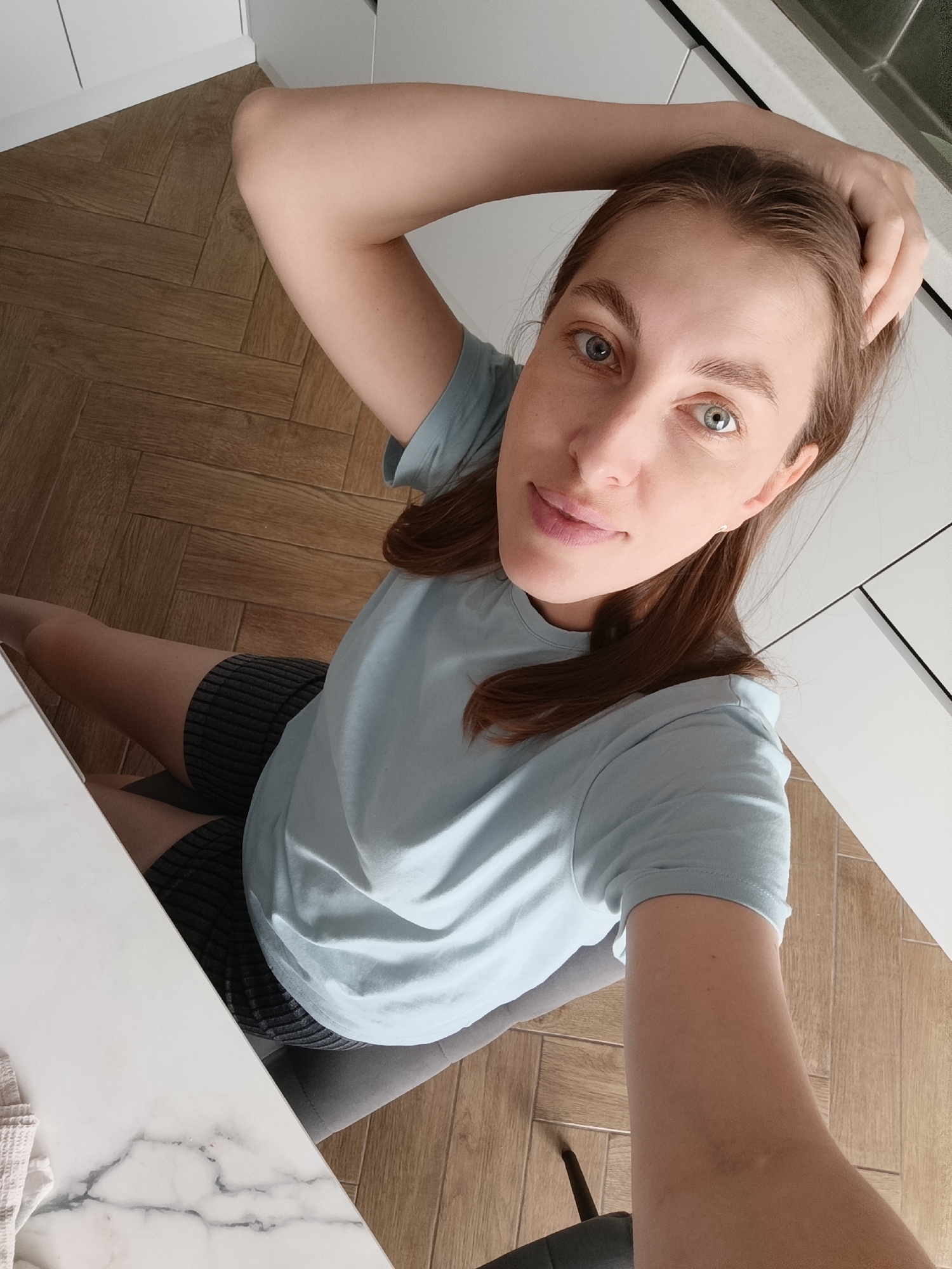 A young woman is captured in a high-angle, close-up selfie from a seated position. She is wearing a light blue t-shirt and dark shorts. Her right arm is bent and resting on her head, with her hand touching her hair. She is looking directly at the camera with a relaxed and natural expression, her blue eyes prominent. Her lips are slightly parted, giving the impression of a gentle smile. The background shows a wooden floor laid in a herringbone pattern, typical of a domestic setting. To the right, white kitchen cabinets are partially visible. The lighting suggests it's daytime, likely indoors, with natural light contributing to the scene. The overall mood is casual and intimate, as if she's taking a moment to herself at home. The image does not contain any visible text.
