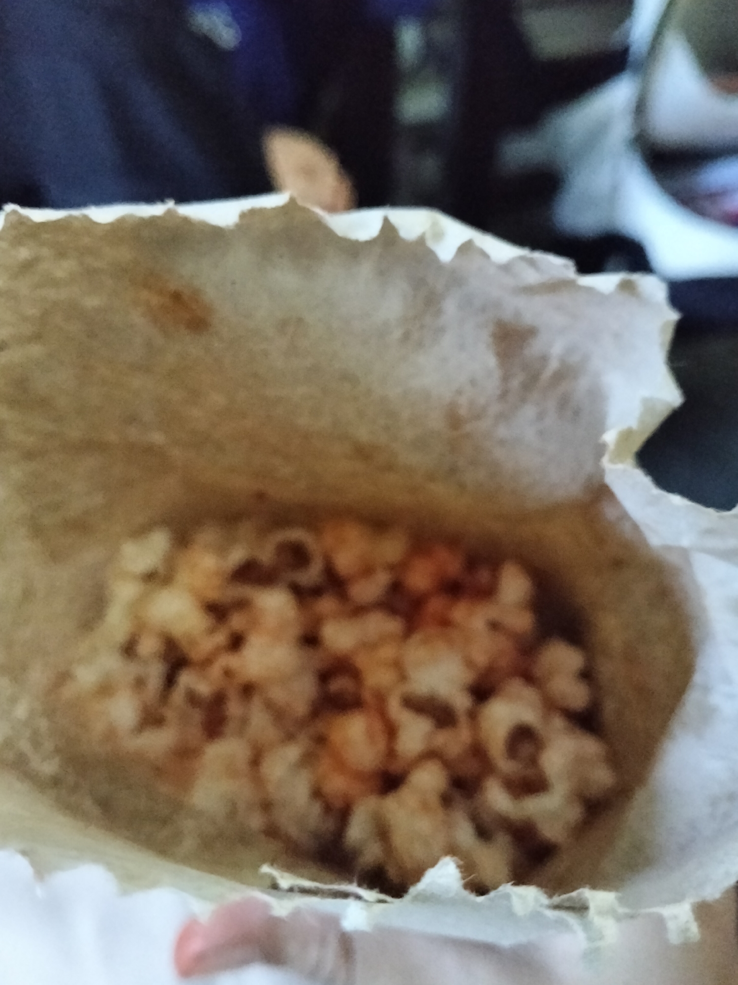 An image displays a close-up of popcorn in a paper bag. The popcorn appears to be seasoned with a reddish spice. The background is blurred but suggests an indoor setting with dark blue and black hues, possibly indicating clothing or furniture. There is no specific indication of the location within Davao City, Philippines.
