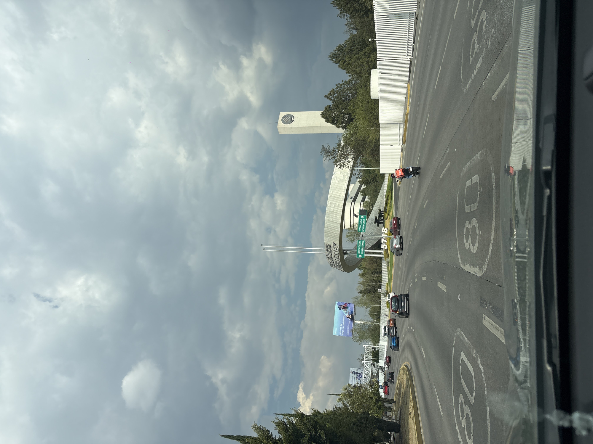 The image shows a highway in Puebla, Mexico. A tall, white tower with a circular logo stands near a curved structure with signage indicating "TECNOLOGICO DE MONTERREY". The sky is overcast with heavy clouds. Cars and motorcycles are traveling on the highway, where speed limits are marked. A billboard is also visible on the side of the road. The scene is taken from inside a vehicle, looking out towards the highway.