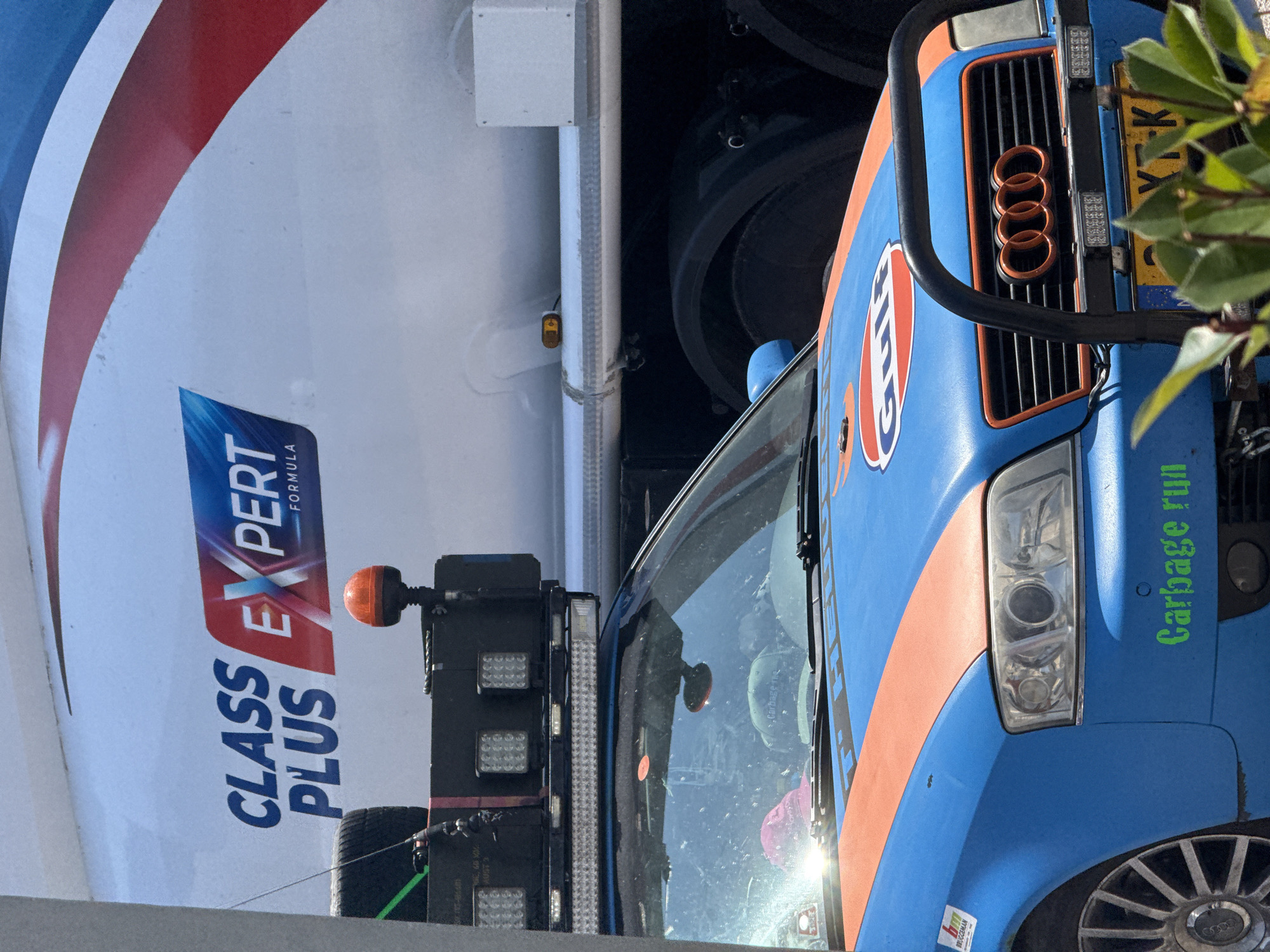 The image shows a close-up view of a blue and orange Gulf-branded vehicle, possibly a truck or van, parked next to a white vehicle with red and blue stripes. The white vehicle has "CLASS EXPERT FORMULA" written on its side. The Gulf vehicle has the iconic Gulf logo and "Gulf" written in white within an orange circle. It also has a grille with four orange rings, resembling the Audi logo.
The front of the Gulf vehicle is visible, including its headlights and a license plate that reads "Y-K". The vehicle appears to be custom-painted with racing stripes. The side of the vehicle has green text that reads "Garbage run". There are also some lights attached to the front of the Gulf vehicle.
The scene appears to be outdoors, possibly in a parking lot or industrial area, given the presence of the two vehicles and the lighting equipment. The lighting suggests it is daytime, and the weather appears to be clear. There are some green leaves from a plant obscuring the top right corner of the image. The image is taken from a low angle, looking up at the vehicles. The overall impression is of a commercial or service vehicle.
The location context is Liješnje, Montenegro.