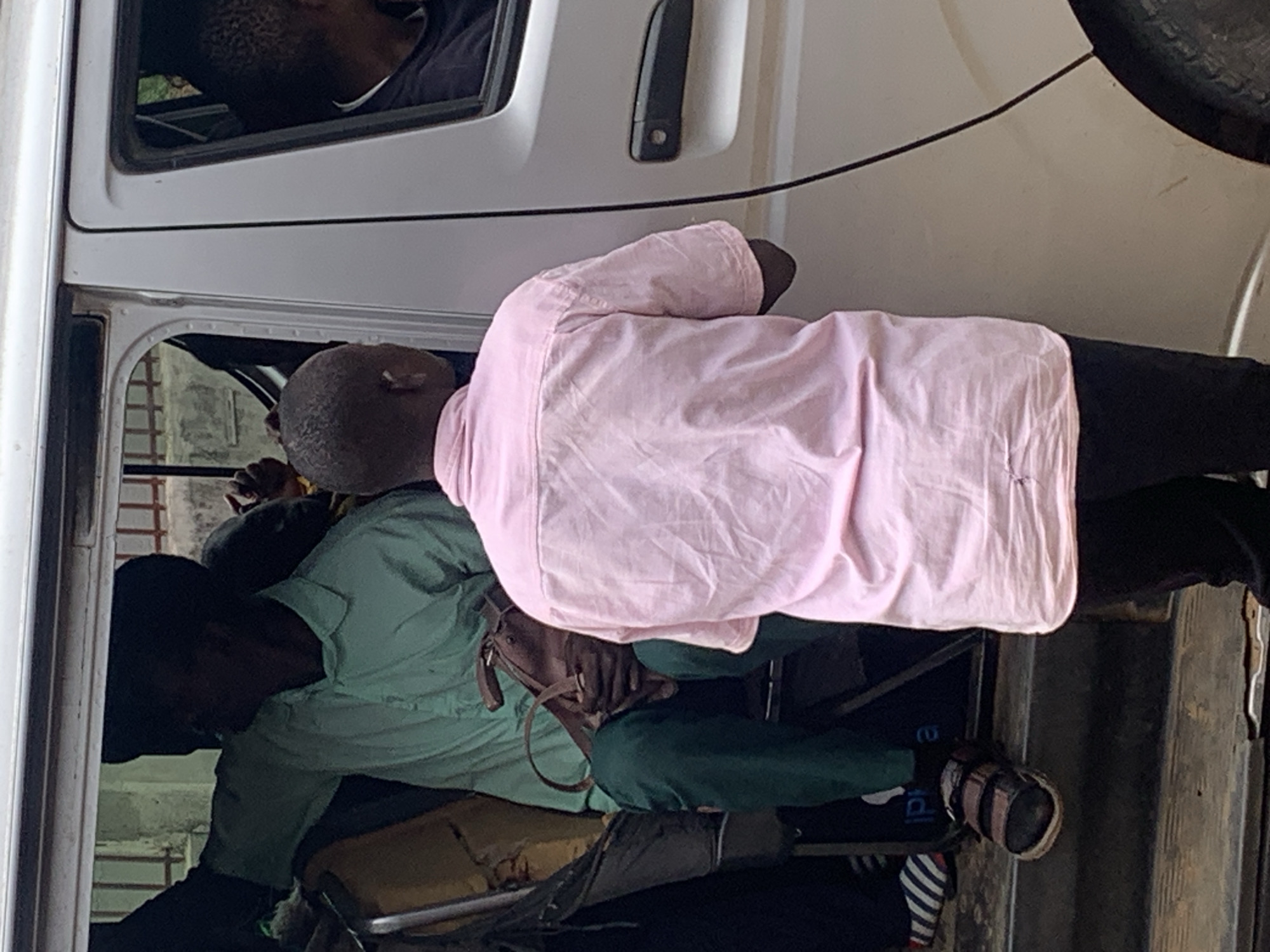 A group of individuals are visible in and around the open doorway of a light-colored van, suggesting it might be a mode of transport. One person, facing away from the camera and wearing a pink collared shirt and black trousers, stands on the step of the van. Another person, in a light green collared shirt and green trousers, is seated inside the van, holding a brown bag. This person is partially obscured by the person standing in the doorway. Additional people are visible within the van. The van appears to be parked outdoors. The environment outside the van is blurred but suggests an urban setting, potentially on a street in Jos, Nigeria. The scene captures a moment of boarding or alighting the vehicle.