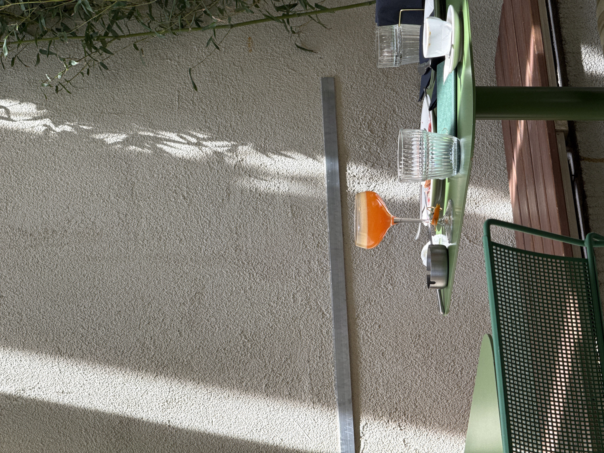 A top-down shot shows a green-topped table set for a meal, outside on a textured, light gray surface, possibly a patio or terrace. The table holds glassware including a tall, ribbed glass and an orange cocktail in a stemmed glass. Dishes and flatware are also on the table. Beside the table, partially visible, is a brown wood slat panel and a green metal chair with a mesh back. To the left of the table runs a metal bar. Light and shadows fall on the wall, indicating the presence of foliage overhead. The location is likely Belgrade, Serbia, based on the visual style of the setting. There are no people visible. The scene captures a moment of outdoor dining in a shaded environment.
