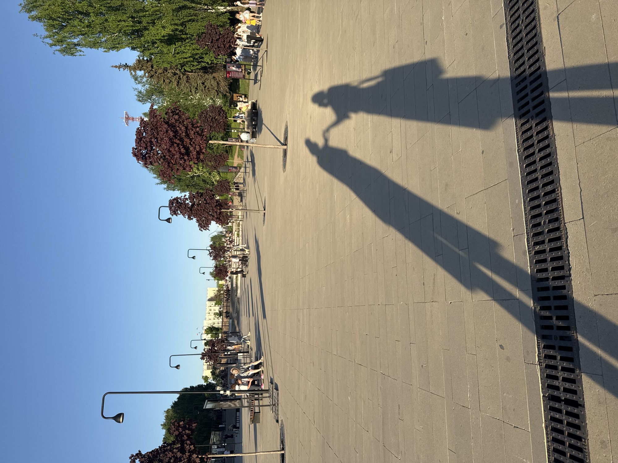 The image shows a long, paved walkway in Izhevsk, Russia, likely a pedestrian zone. The walkway is lined with young trees with reddish-purple foliage, interspersed with modern street lamps with curved arms. Benches are positioned along the walkway where people can be seen sitting. In the distance are buildings, indicating an urban environment. The sky is clear and blue. The scene is illuminated by strong sunlight, casting long shadows. The shadows appear to be of two people, one possibly pushing the other in a wheelchair or stroller. A metal drainage grate runs along the edge of the walkway. A tall lattice tower can be seen in the far distance.