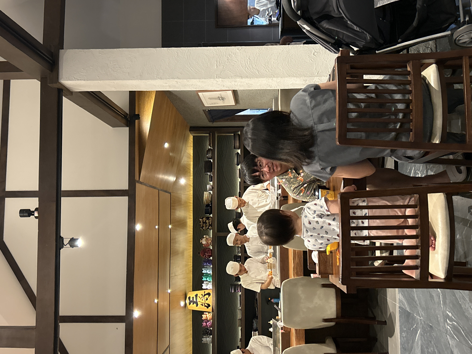The interior of a restaurant in San Sai Luang, Thailand, shows a sushi bar with several chefs wearing white uniforms and hats behind the counter. A family is seated at the bar; a woman with dark hair and a gray shirt is seated in a wooden chair, with a child in a patterned shirt beside her. Another person is partially visible next to her, wearing glasses and a patterned shirt. A stroller is in the background near the ceiling. The interior design features wooden beams, white walls, and warm lighting. A display of colorful items is seen behind the sushi chefs, and a lit sign with characters is visible. The floor appears to be marble or a similarly patterned material.