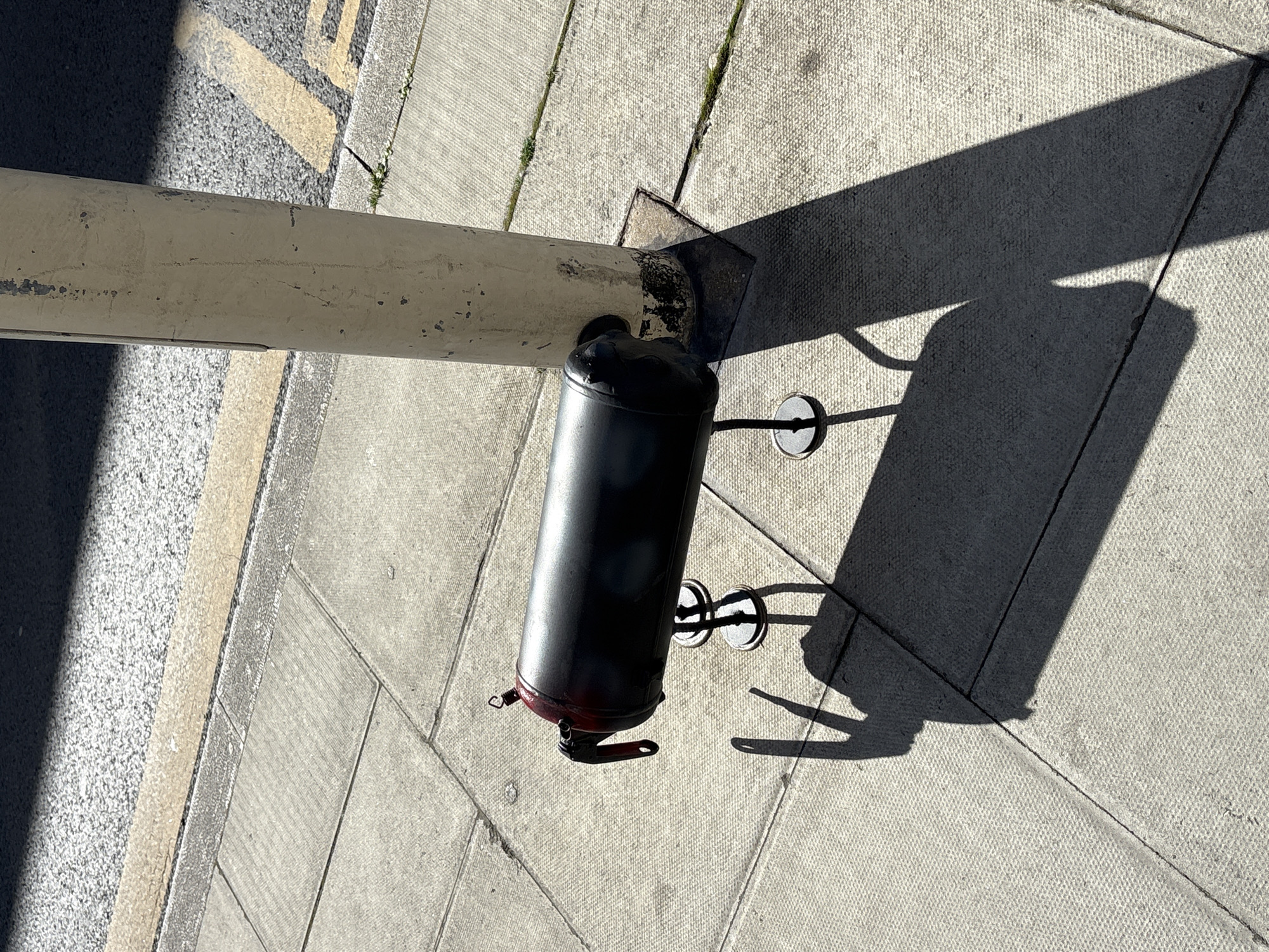 An eye-level, outdoor photograph shows a black, cylindrical object attached to a pole and mounted on a paved street surface. The pole, oriented horizontally, is painted a weathered cream color and appears to be metal. It is attached to a dark, metal base plate bolted to the paved surface. The black cylinder is affixed to the base plate. It has two, thin, metal rods extending outwards. These rods each terminate in a circular, flat piece. The object casts a sharp, dark shadow on the paving stones. The paving is light gray and formed from rectangular blocks. In the background, part of a curb and street surface is visible, featuring yellow markings. The scene appears to be within a city in London, United Kingdom.