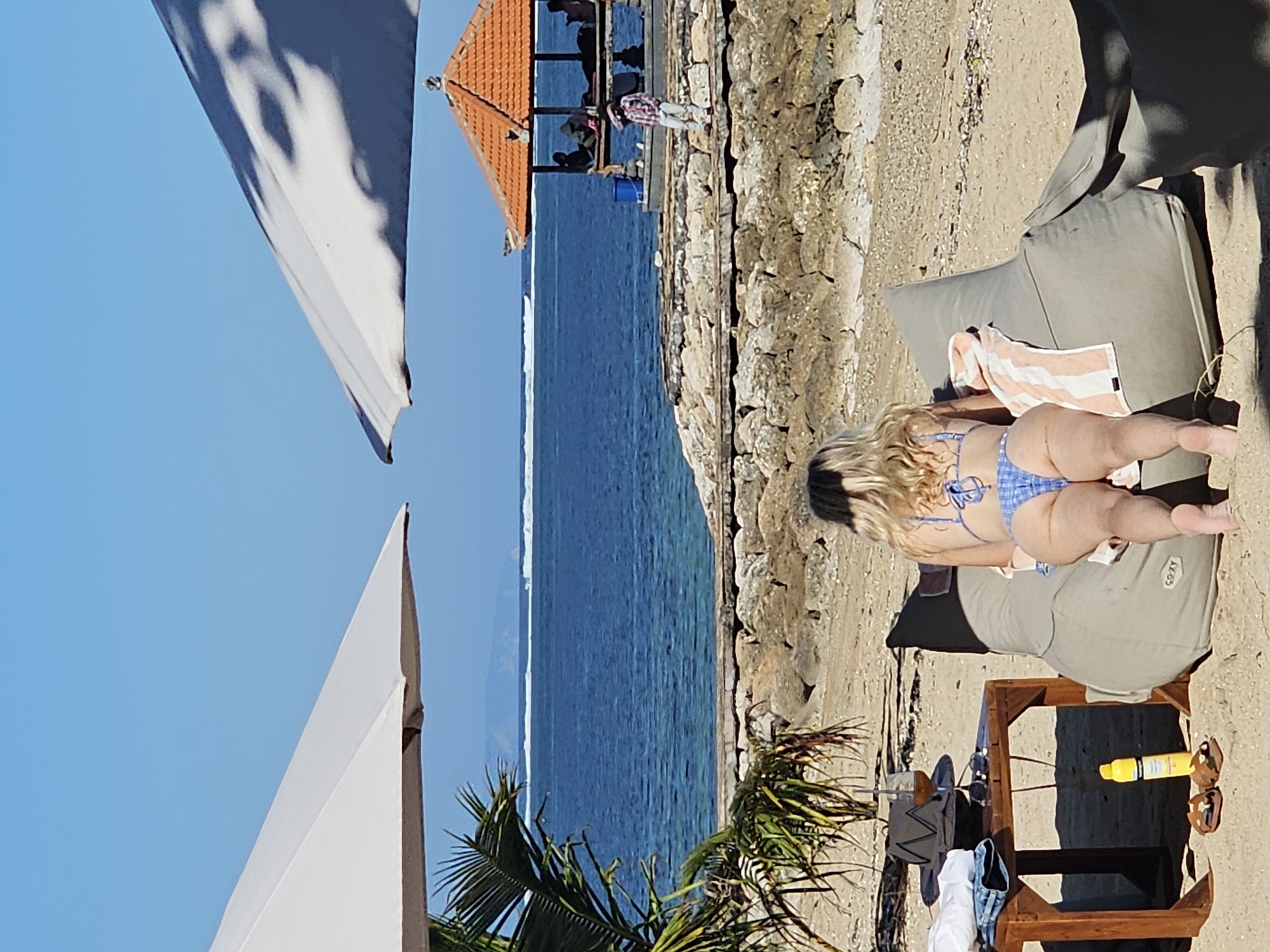 The image depicts a sunny day at a beach in Denpasar, Indonesia. In the foreground, a woman wearing a blue and white checkered bikini is lying on a large, gray beanbag chair on the sand. Her back is to the camera, and she is facing the ocean. To the left of the beanbag, there is a small wooden table with a glass of drink, a hat, sunglasses, and folded clothes. A bottle of sunscreen and a pair of sandals are on the sand next to the table.
Beyond the beach, a stone wall separates the sand from the ocean. The blue water has gentle waves, and in the distance, a line of white surf indicates a reef. Further out, a hazy coastline with mountains can be seen. On a rocky outcrop in the middle ground, there is a traditional Balinese-style open-air pavilion with an orange tiled roof, where several people are seated. A person in a red and white checkered shirt is walking on the rocks near the pavilion. Two large white beach umbrellas are positioned in the scene, one on the left and one on the right, casting shadows on the sand and the water. A palm tree frond is visible on the left side of the image. The sky is a clear, bright blue.