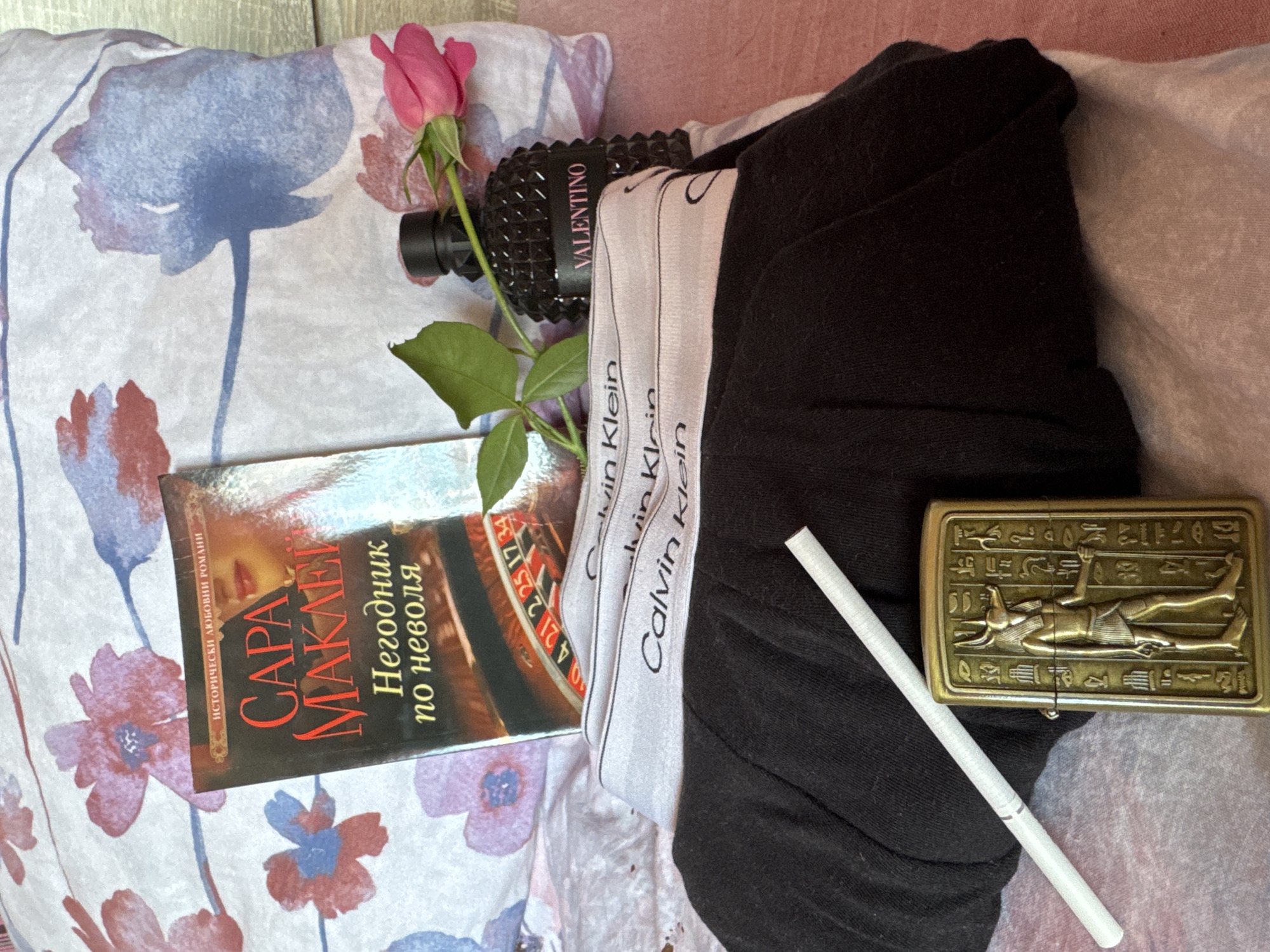 On a bed with floral patterned bedding, a book titled "CAPA MAKEIN Негодник по неволя" is positioned next to a pink rose and a black Valentino perfume bottle. A pair of black Calvin Klein underwear is folded nearby, along with an Egyptian-themed lighter and a white cigarette. The scene is set in Dolna Oryahovitsa, Bulgaria.