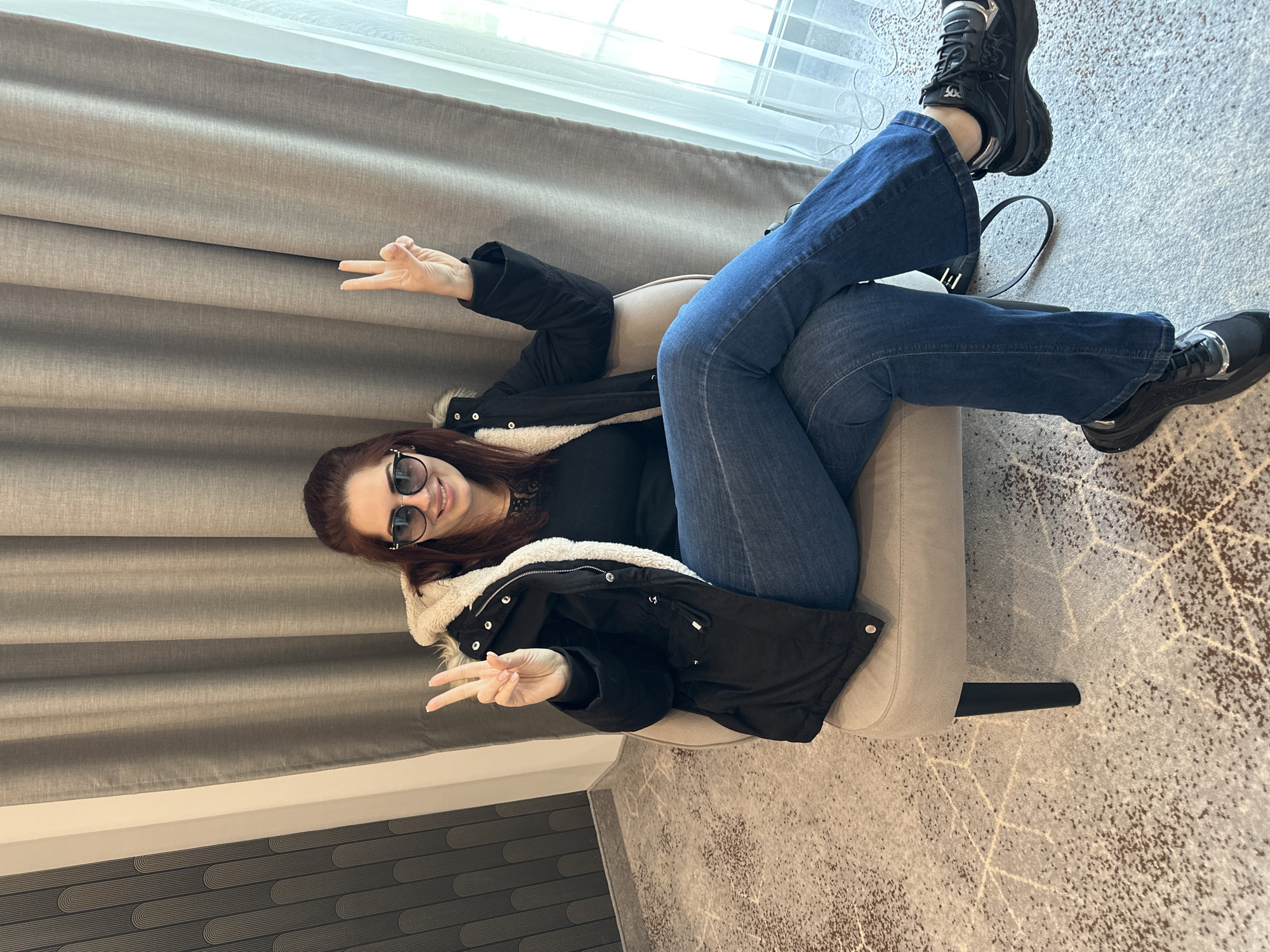 A woman with shoulder-length auburn hair and sunglasses is reclining in a light-beige armchair. She is wearing blue jeans, a black top, and a black jacket with a cream-colored fleece lining. Her legs are crossed, and she is making the peace sign with both hands. She is in a room with light-gray curtains, a window, and patterned carpeting. The location is not discernible from the image.