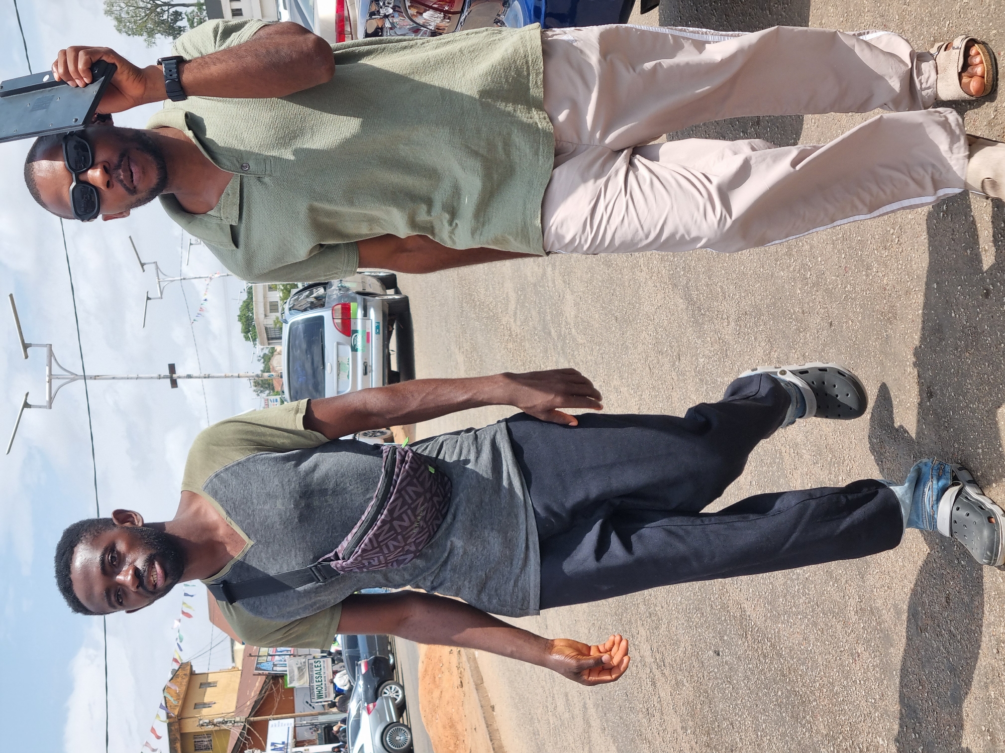 Two African men stand outdoors. One wears a green shirt, khaki pants, sandals, sunglasses, and a wristwatch, holding a black tablet. The other wears a gray and green shirt, black pants, Crocs, and a purple patterned fanny pack. Behind them, cars are parked on the street. The location is Jos, Nigeria.