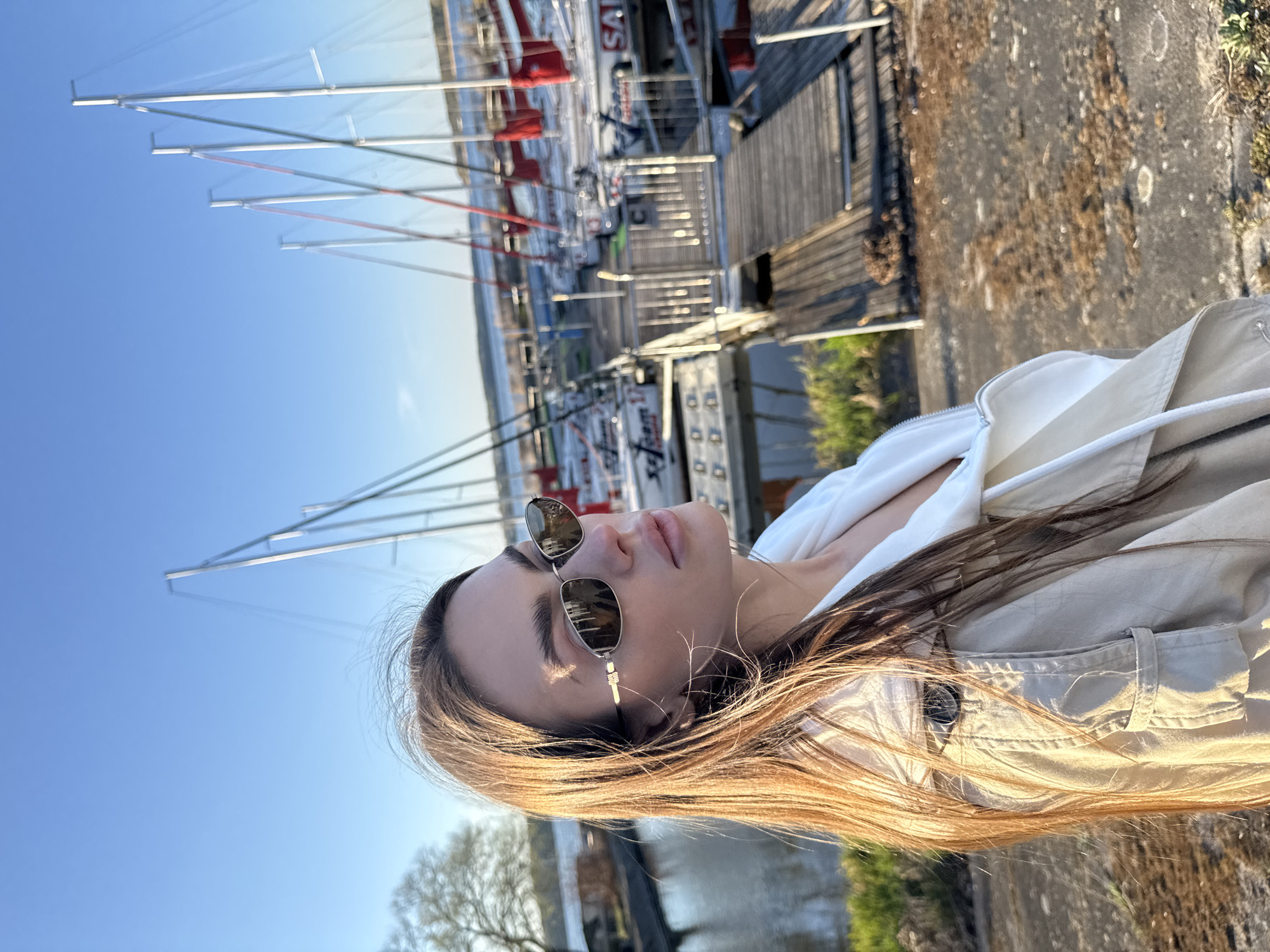A young woman with long brown hair and sunglasses is standing on a concrete surface near a body of water, possibly a river or harbor, in Białobrzegi, Poland. In the background, there are several sailboats docked at a pier. The sky is blue and clear. The woman is wearing a white hoodie underneath a light beige jacket. The area appears to be a marina or waterfront area.
