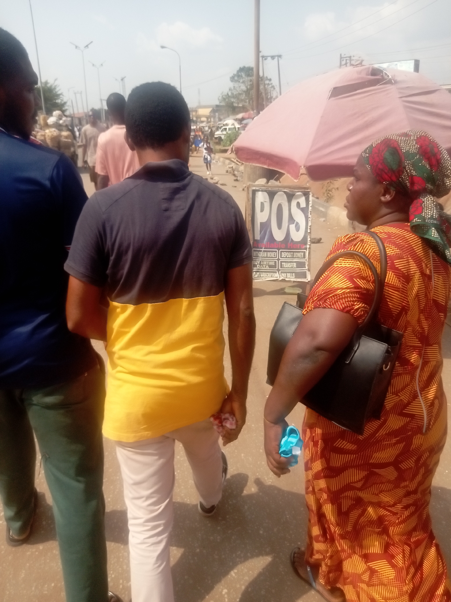 The image depicts a bustling street scene in Onitsha, Nigeria, under clear, bright daylight. In the foreground, three individuals are partially visible, generally moving towards the left. On the right, a woman with dark skin is seen in profile, looking left. She wears a patterned long dress with orange, red, and yellow geometric lines, and a matching headwrap. A black shoulder bag hangs on her right shoulder, and she holds a small, light-blue object in her left hand. To her left, slightly ahead, a man is viewed from behind, wearing a short-sleeved polo shirt that is dark gray on top and yellow below, with light-colored trousers and dark shoes. Further left and partially obscured, another man is seen from behind, wearing a dark blue short-sleeved shirt and green trousers.
In the mid-ground, behind the woman, a large, weathered, light-pink market umbrella provides shade. Beneath the umbrella, a white sign with "POS" prominently displayed in black letters advertises financial services including "Withdraw Money," "Deposit Money," "Buy Airtime," "Transfer," "Pay Subscription," and "Pay Bills." The background reveals a busy street or market area with several individuals walking in the distance, some carrying goods. Overhead, multiple streetlights with dark poles are visible, along with power lines. Buildings and vehicles are indistinctly visible further down the street. The ground is an uneven, light-colored surface, possibly concrete or dirt, suggesting a well-trafficked urban pathway.
