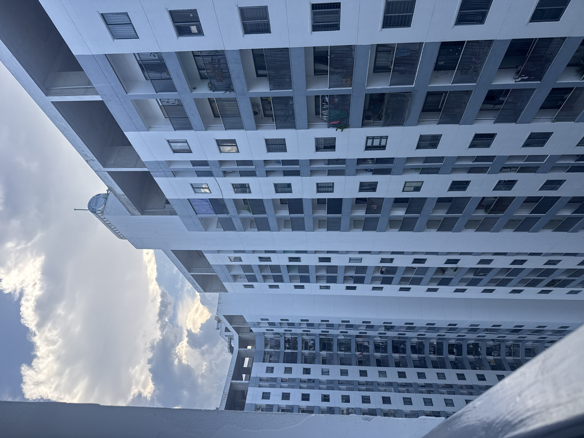 The image shows a tall, modern apartment building in Nha Trang, Vietnam, with a cloudy sky above. The building has a repetitive facade with many balconies and windows. The architecture is contemporary, with a mix of white and gray surfaces. The balconies have dark railings and some appear to be furnished with outdoor seating or plants. Some balconies have clothes hanging out to dry, suggesting the building is inhabited. The overall impression is of a densely populated urban environment.