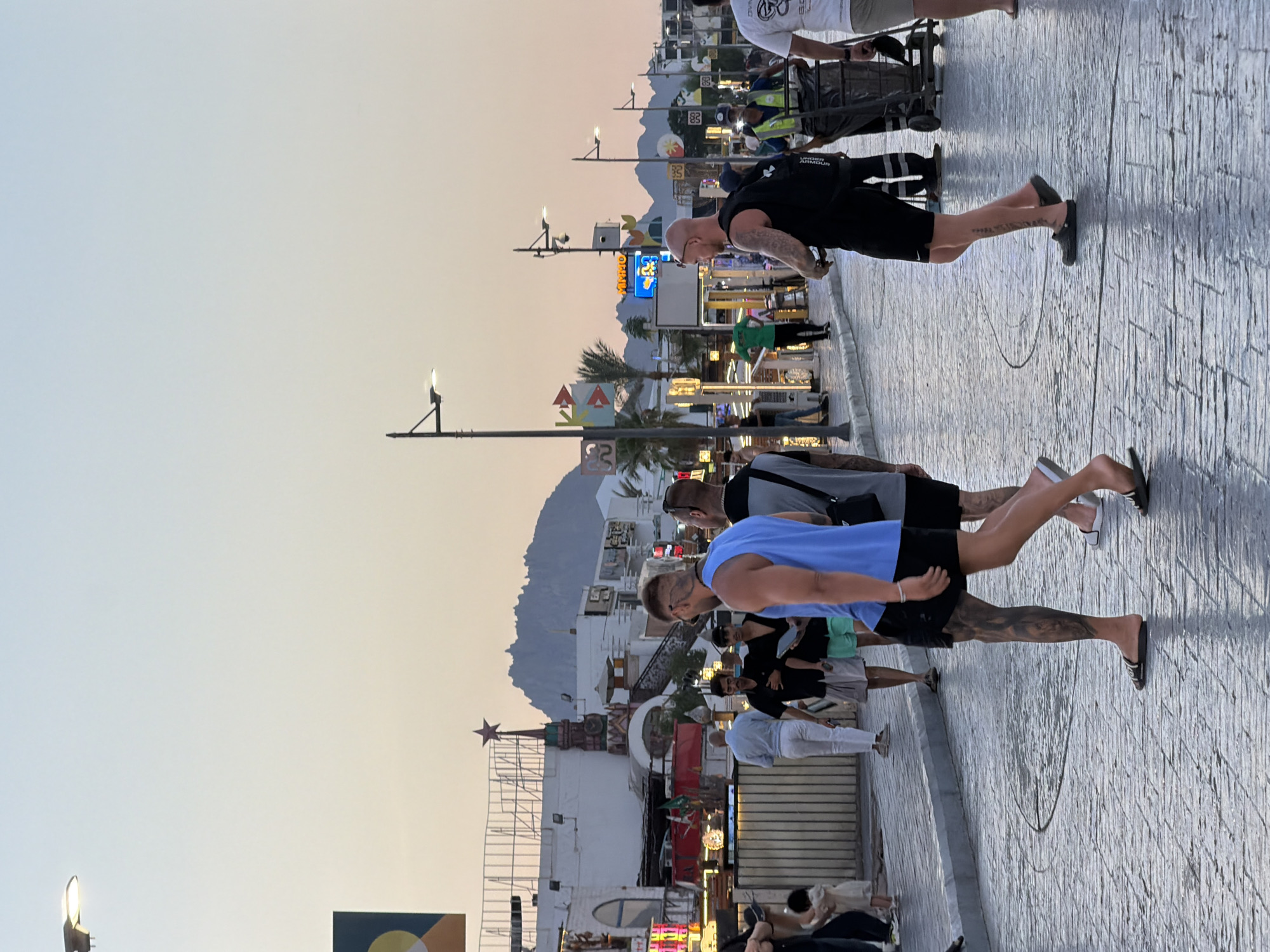 The image depicts a street scene in Sharm El Sheikh, Egypt, during what appears to be late afternoon or early evening. The sky is a soft gradient of pale blue to orange, suggesting a sunset. The street is paved with textured cobblestones that reflect the ambient light.
Several people are walking along the street. In the foreground, three men are walking towards the right. The man closest to the camera is wearing a blue tank top and black shorts, with extensive tattoos on his legs and arms. He is wearing black flip-flops. To his left, another man in a grey shirt and black shorts is also walking. Further to the right, a bald man in a black tank top and black shorts is walking, carrying a black backpack. He is wearing dark clogs.
In the background, there are various buildings with storefronts and signage, illuminated by colorful neon lights. Some of these signs are in English. A distinctive building with a red star on top, resembling a tower, is visible. Palm trees and other vegetation can be seen interspersed between the buildings. Further in the distance, a mountain range is silhouetted against the sky.
Other individuals are also visible in the midground and background, including a woman in a white dress, a child, and individuals appearing to be street vendors or workers, one of whom is pushing a cart with bins. Streetlights with modern designs are mounted on poles along the street. The overall atmosphere suggests a busy, tourist-oriented commercial area.