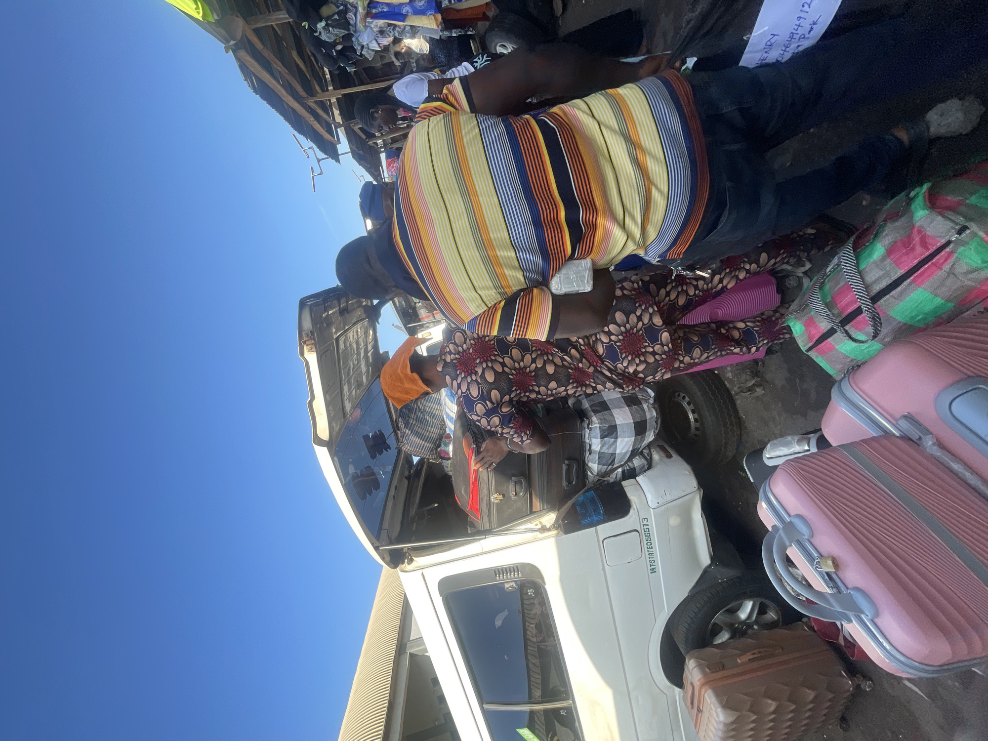 In Ado-Ekiti, Nigeria, at what appears to be a bus park, a white van is being loaded with luggage. A man in a striped shirt and jeans is standing beside the van, holding a clear plastic bag. A woman in a patterned dress is placing items into the van. Several suitcases, including pink and brown ones, are on the ground near the van. In the background, market stalls with various goods are visible under a corrugated metal roof. The sky is clear and blue. A sign on the side of the van reads "INTSTATE058573". Another sign on a piece of paper near the man states "HENRY 0461944912 + Park".
