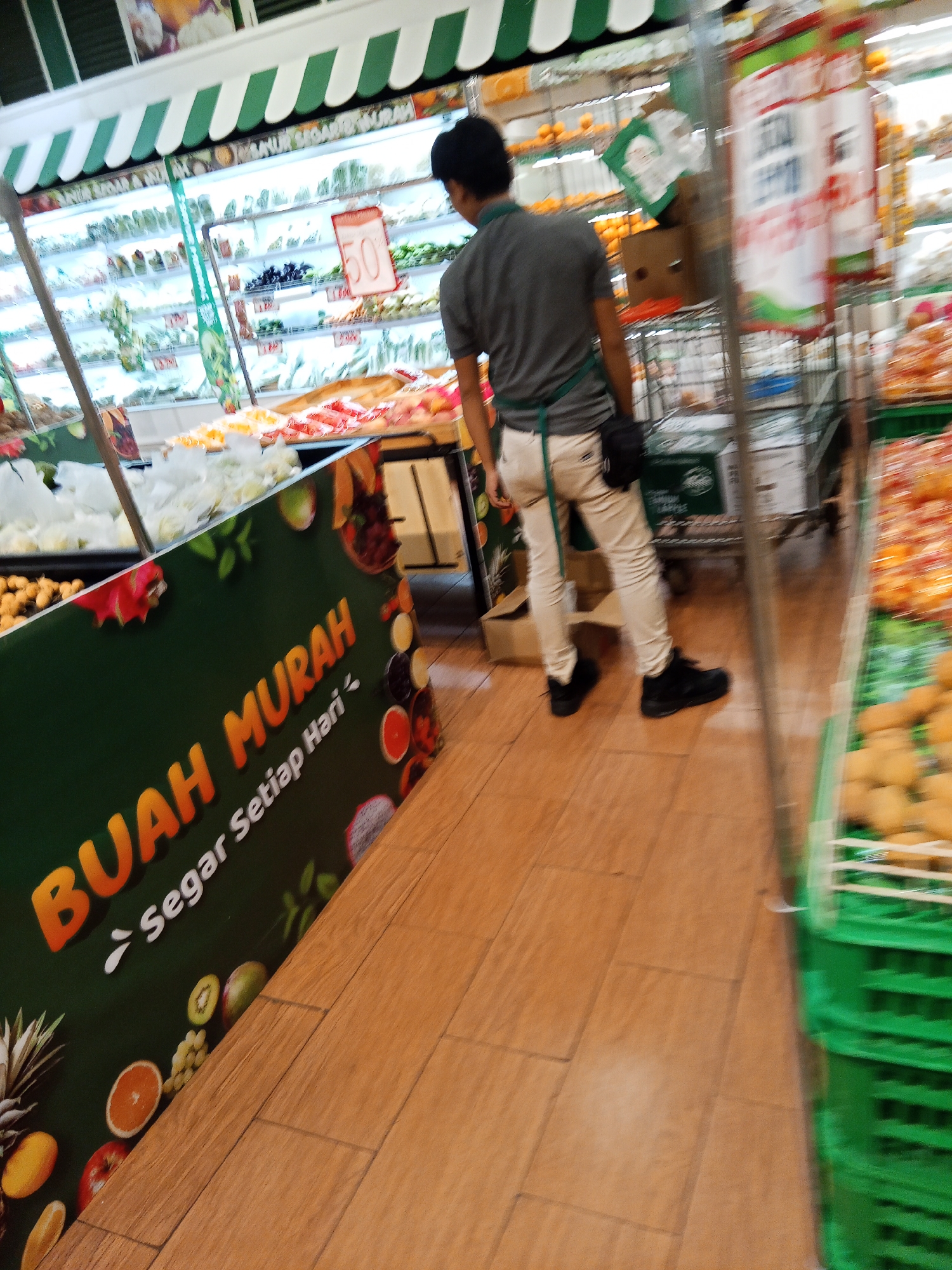 The image captures a scene inside a supermarket in Lubuklinggau, Indonesia. A man, wearing a gray t-shirt, beige pants, and black shoes, is standing in the aisle. He is seen from the side, with his back mostly towards the camera. He appears to be wearing a green apron or sash. The supermarket aisle is stocked with various fruits and vegetables displayed on shelves and in crates. In the foreground, a large green banner with orange lettering reads "BUAH MURAH" (Cheap Fruits) and "Segar Setiap Hari" (Fresh Every Day). The floor is tiled with wooden planks. Other shoppers or supermarket staff are not clearly visible. The lighting is bright, typical of an indoor retail environment.