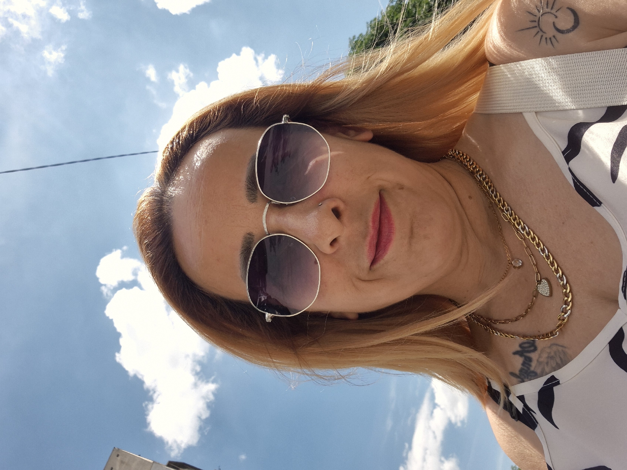 In this close-up, overhead selfie, a woman with blonde hair and sunglasses poses outdoors under a bright blue sky dotted with white clouds. She is wearing a white top with a black abstract pattern and multiple necklaces, including a gold chain with a heart-shaped pendant. A tattoo of a sun and crescent moon is visible on her right shoulder. The background suggests an urban environment with hints of buildings and trees.
