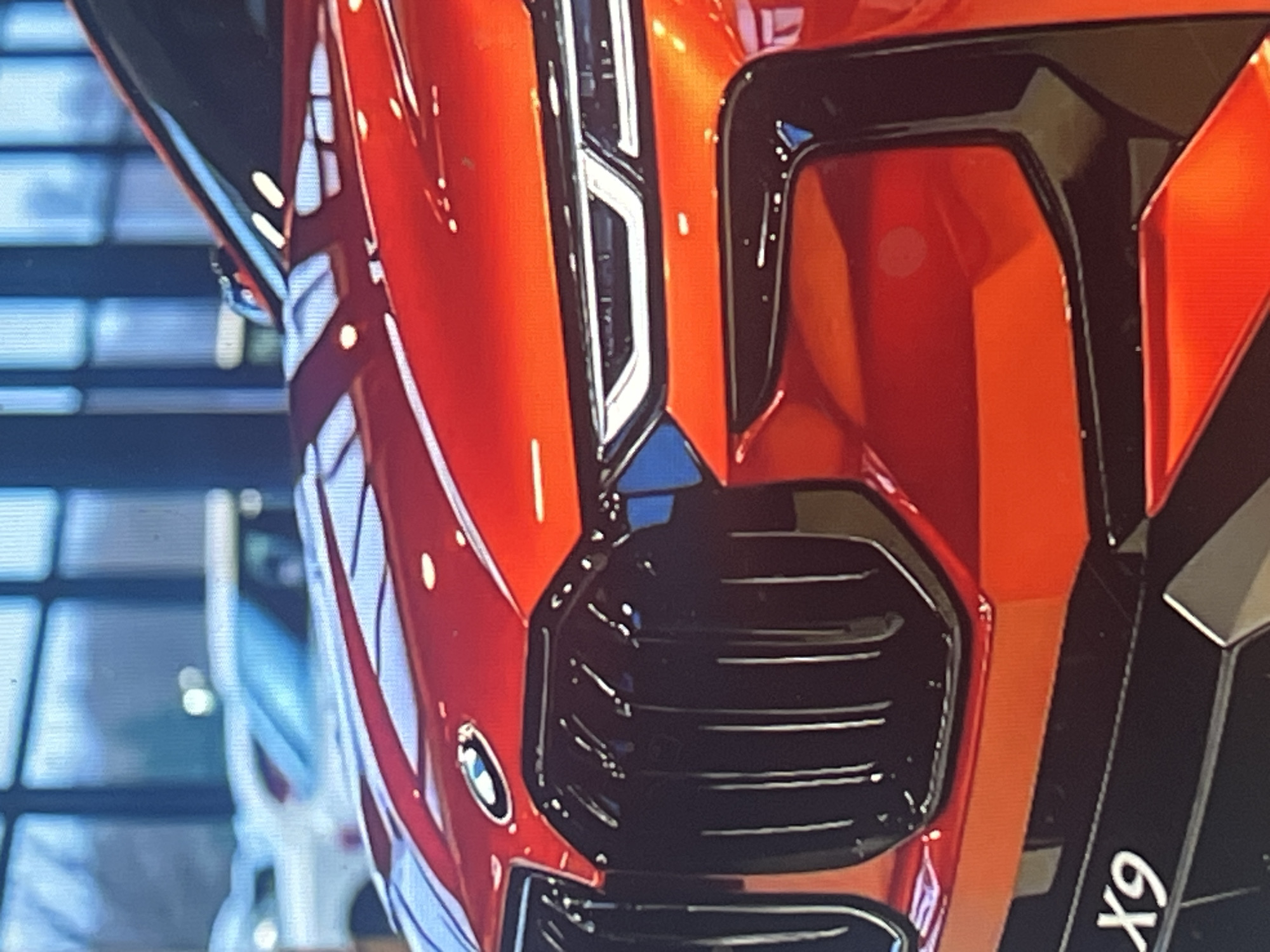 This is a close-up, slightly blurry, eye-level shot of the front of a red car, likely a BMW, as indicated by a visible BMW logo. The car is shiny, reflecting light sources. The focus is on the front grille and headlights. The grille is black and features horizontal slats. The headlights are sleek and angular, with white and black accents. The car's bodywork is a vibrant, glossy red. To the left of the grille, there are some diagonal white stripes with blue outlines, possibly part of a pattern or decal on the car.
In the background, out of focus, are what appear to be windows or a tiled wall with a grid pattern. The overall setting seems to be indoors, perhaps a showroom or a garage, given the clean environment and the lighting. There are no people visible in the image. The activity appears to be a static display of the car. The visible text "X9" is on a black surface on the right side of the image, oriented vertically and in white lettering. The time of day and weather are not discernible. The location context of Vanadzor, Armenia, is provided externally.