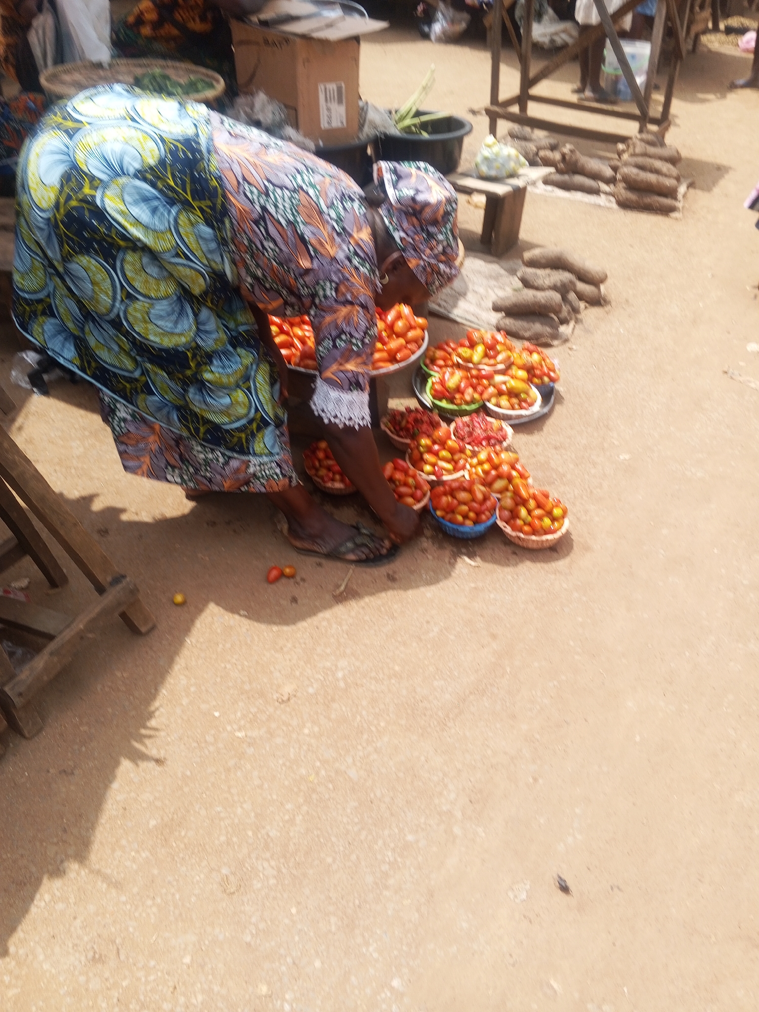 This image captures a market scene in Kabba, Nigeria. A woman, wearing a vibrantly patterned blue, yellow, and orange traditional attire, is bent over a selection of fresh tomatoes displayed in various bowls and baskets on the ground. The tomatoes are predominantly red and yellow, indicating ripeness. Behind the woman, several stacks of yams, a staple food in the region, are visible, along with other market goods and structures. The ground is a sandy, earthy color, typical of an outdoor market. The overall impression is one of a bustling local marketplace where fresh produce is being sold.