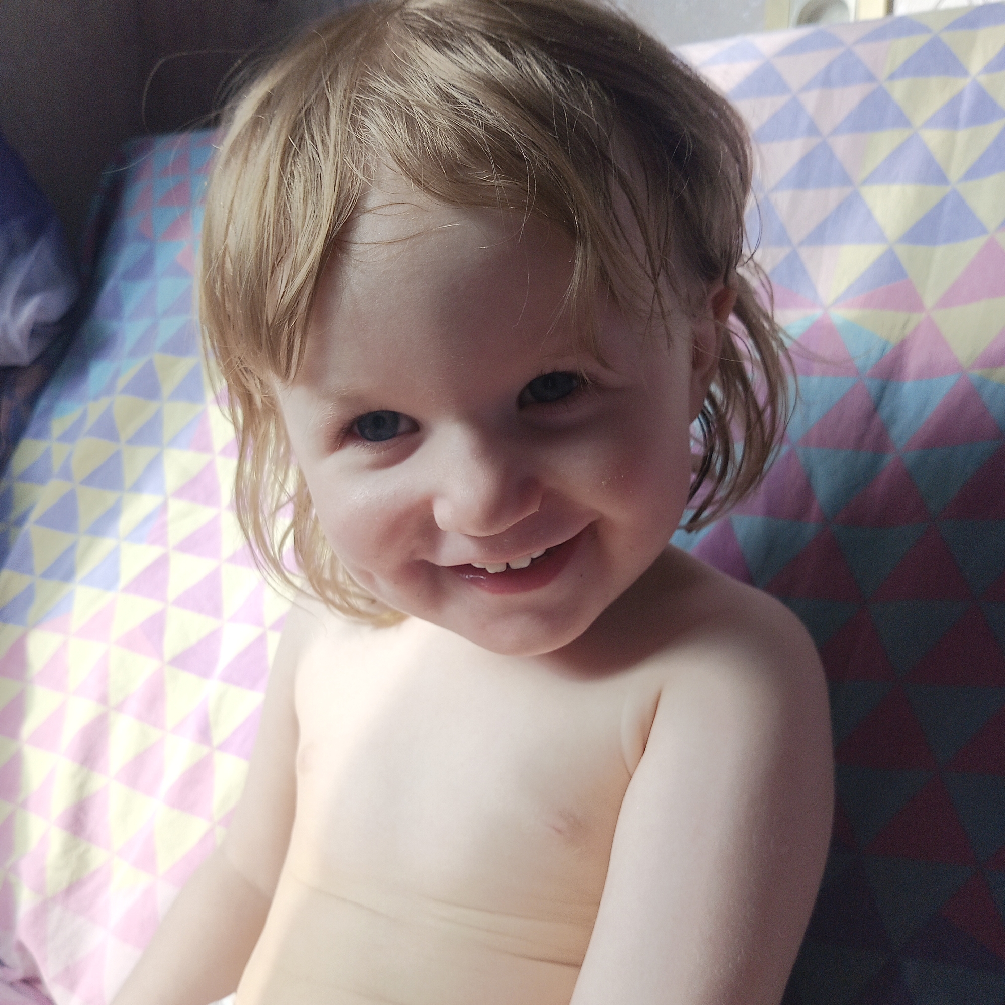 A young, fair-skinned child, likely a toddler, is captured in a close-up portrait, smiling widely at the camera. The child has light blonde or strawberry blonde hair styled with a short, shaggy cut, and bright blue eyes. Their joyful expression is evident with a broad smile that reveals several small teeth. The child appears to be shirtless or wearing minimal clothing on their upper body.
The background consists of a brightly colored fabric, possibly a bedsheet, pillowcase, or blanket, adorned with a geometric pattern of numerous triangles. These triangles are rendered in various pastel and slightly darker shades, including pink, light yellow, light blue, purple, and some darker blue and reddish-brown hues, forming a quilt-like design. The lighting is soft and natural, suggesting an indoor setting during daytime. No specific activity is taking place other than the child looking and smiling. No visible text is present in the image. This scene takes place in Chaikovsky, Russia.