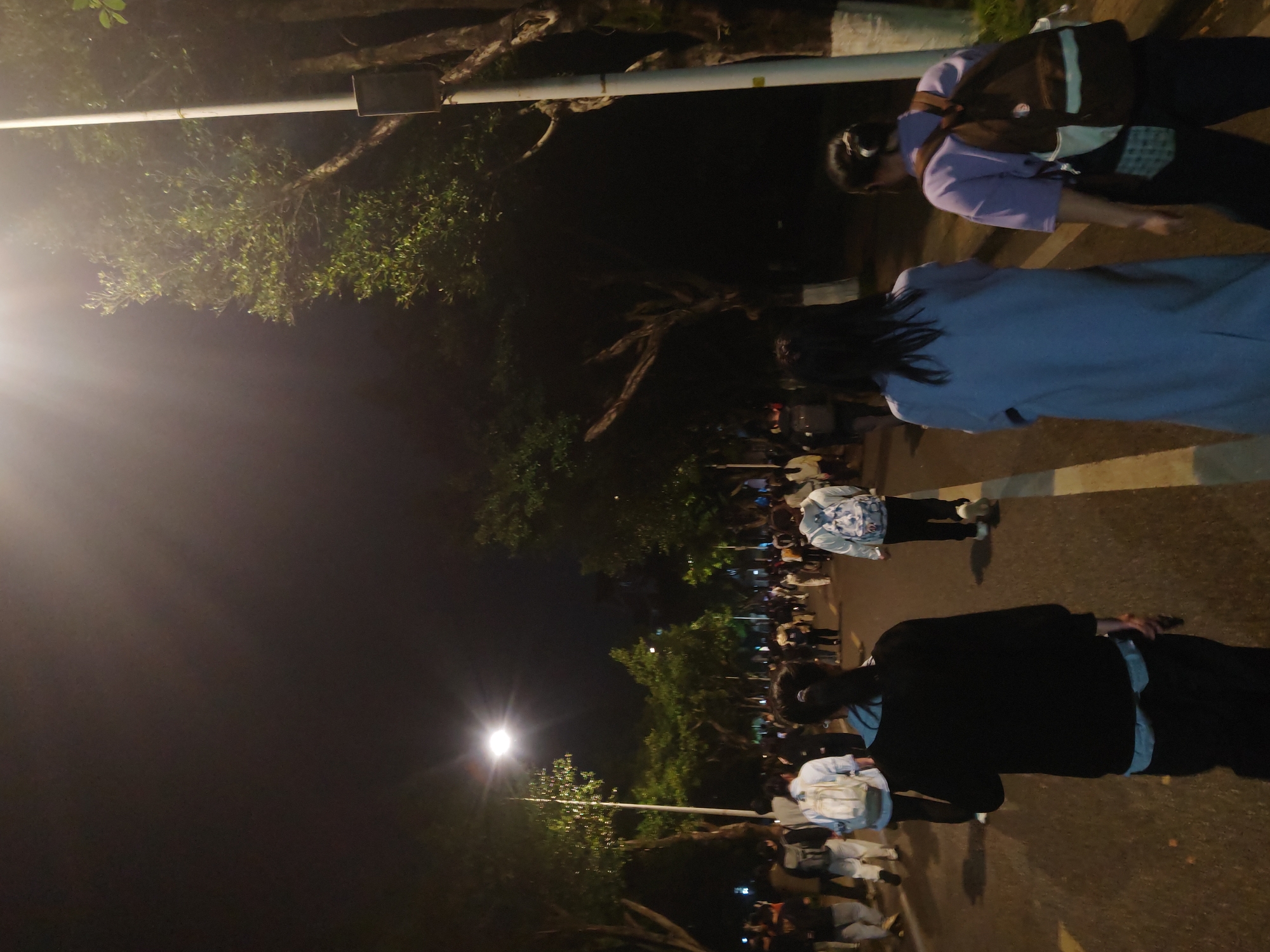 The image shows a crowd of people walking on a street in Chongqing High-tech Zone, China at night. Streetlights illuminate the scene. People of various ages and wearing different clothing are present. Some are walking individually while others are walking in groups. The road surface is paved. Trees line the side of the street.