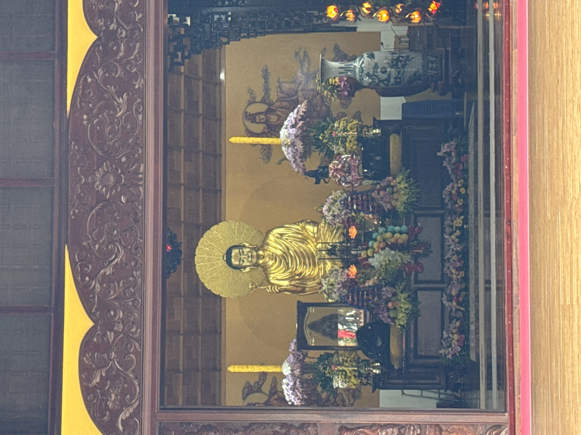The image captures a vibrant and serene interior of what appears to be a Buddhist temple or shrine in Da Lat, Vietnam, viewed through an elaborately carved dark wooden frame.
The central subject is a golden Buddha statue, positioned on a tiered altar. The Buddha is depicted with serene features, a contemplative expression, and a prominent golden halo or aureole behind its head, signifying its spiritual radiance. It wears flowing robes, characteristic of Buddhist iconography.
Surrounding the statue and adorning the altar are numerous colorful offerings, predominantly fresh and artificial flowers in various hues of purple, pink, white, and yellow, arranged in abundant bouquets and garlands. Several rounded, colorful objects resembling fruit or decorative spheres are also part of the offerings. On the right side, a tall, elegant blue and white ceramic vase with intricate patterns holds another floral arrangement. A long, slender yellow object, possibly a large candle or incense stick, stands on the left side of the statue, with another partially visible below on the right. Below the main Buddha, a smaller altar holds a framed mirror, reflecting a glimpse of the surroundings.
The background wall is painted a warm, light yellowish-brown, featuring murals of other spiritual figures or deities, depicted in a traditional style with halos. The ceiling above is visible, showcasing patterned dark wood or decorative panels. The ornate wooden frame in the foreground is intricately carved with swirling floral and organic motifs, providing a sense of grandeur and craftsmanship. Warm, glowing lights, possibly decorative lanterns, are visible at the very top of the scene, adding to the tranquil and spiritual ambiance. The overall impression is one of deep reverence and devotion in a richly decorated sacred space.