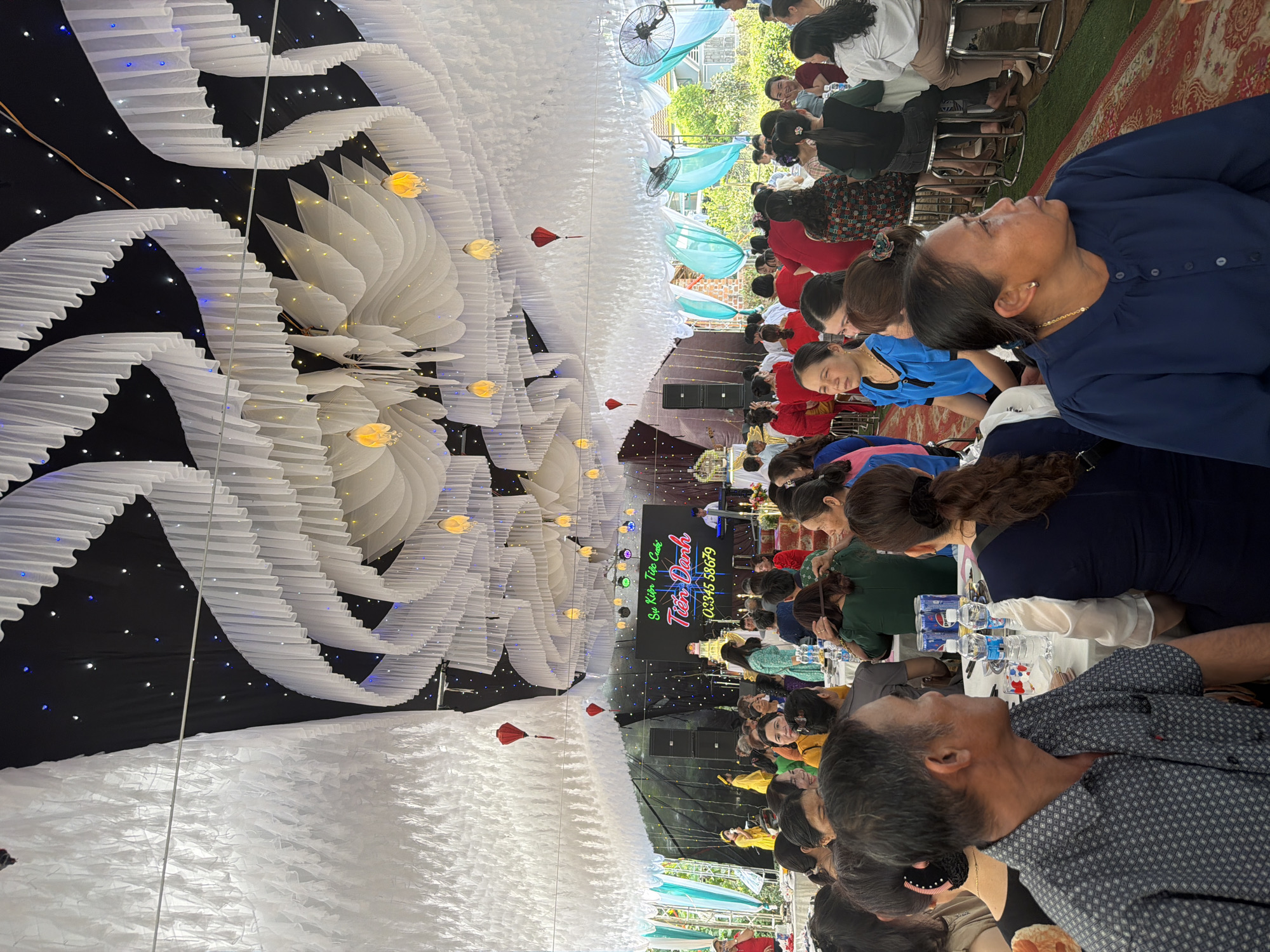 The scene is an outdoor event with many people seated at tables under a large tent-like structure, which is decorated with white fabric and hanging lights. There is signage that says, "Sự Kiện Tiệc Cuối Tuần Danh Tiến Danh" followed by a phone number. People are mostly seated and appear to be eating or talking. The location is likely Bình An, Vietnam.