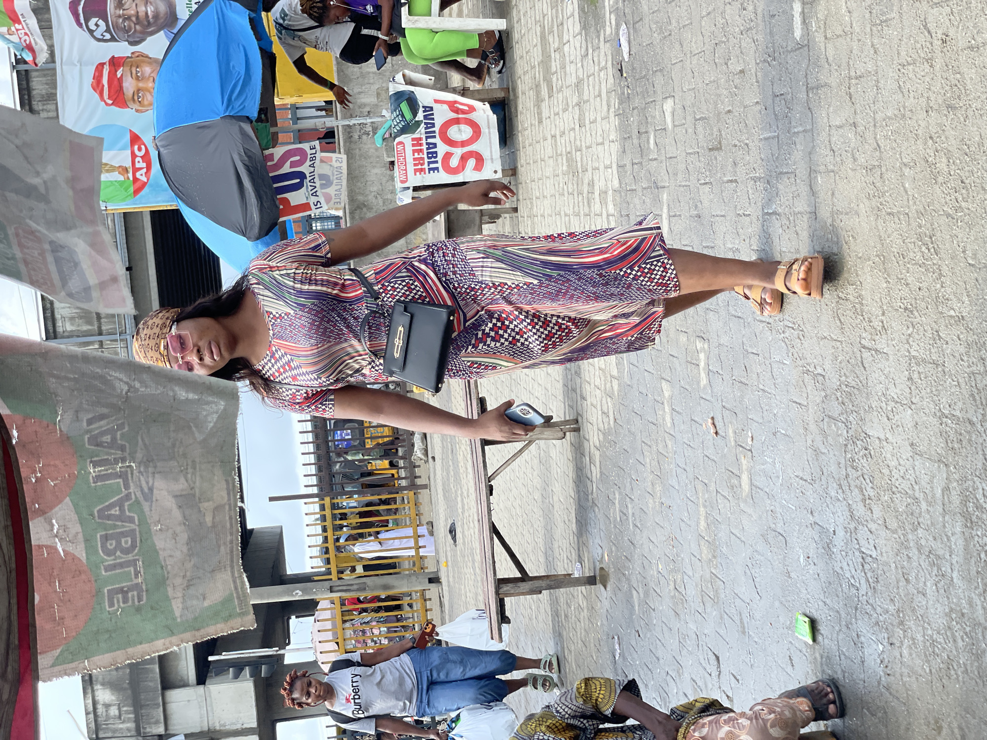 The image captures a street scene in Lekki, Nigeria. The foreground features a woman wearing a patterned dress, sunglasses, and sandals. She is holding a phone and has a black purse. In the background, there are people, political posters, a POS sign, and a covered structure with text. The ground is paved with patterned tiles. The scene includes elements of urban life with commercial and political activity.
