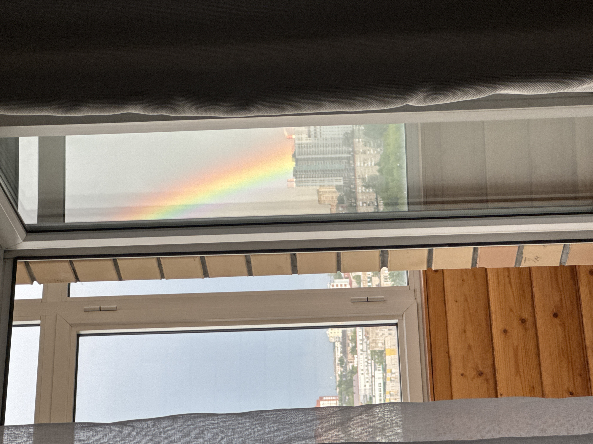 View from a window in Perm, Russia. The scene shows two windows framed by a gray curtain at the top and a partially visible sheer curtain at the bottom. The top window displays an inverted reflection of the cityscape, with a vibrant rainbow arc visible on the left. The lower window shows a more direct view of the city, also upside down. To the right of the lower window, there's a wall covered in light brown wooden paneling. The buildings in the cityscape appear to be residential apartments.